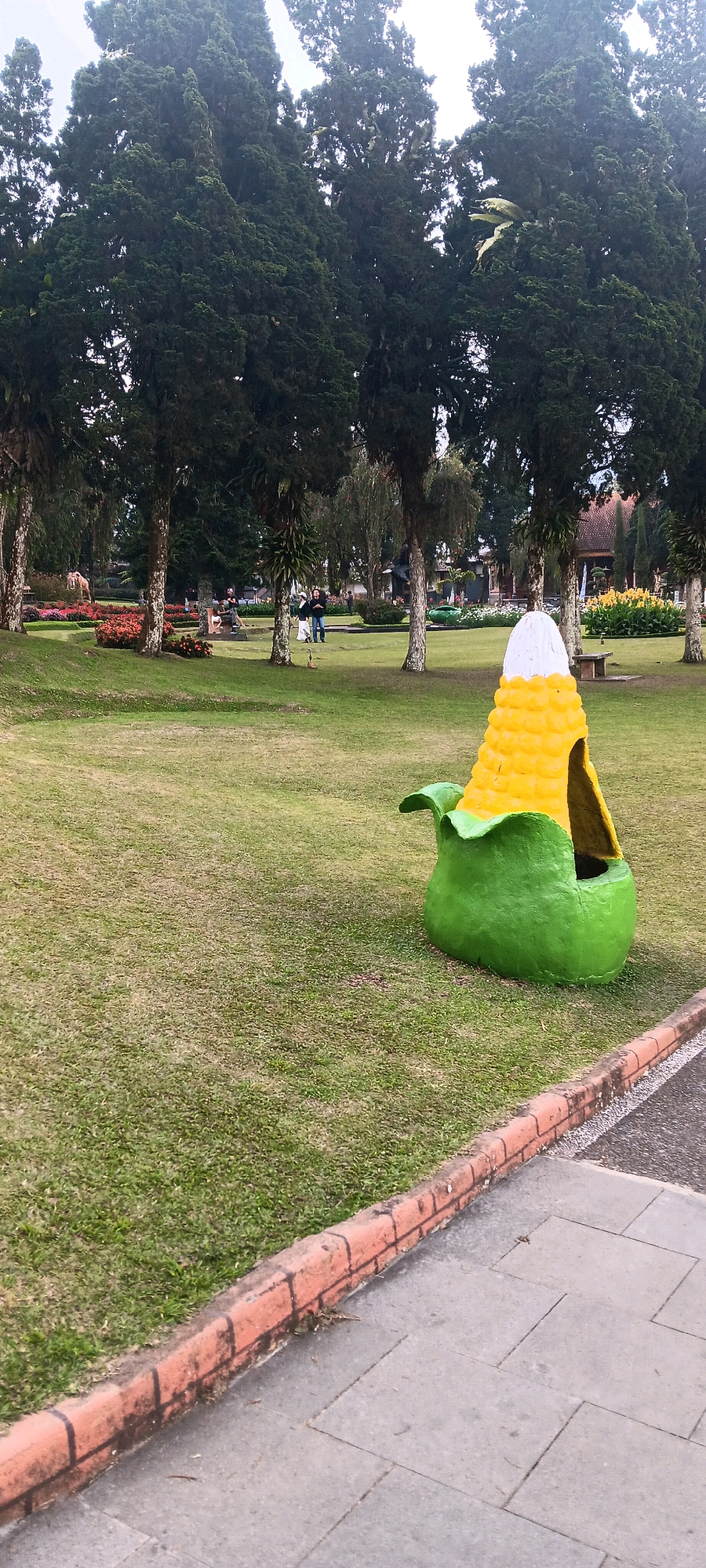 In a grassy park in Baturiti, Indonesia, a whimsical corn-shaped structure with a green base and yellow kernels sits on the lawn. The structure appears to be a playhouse or decorative element, with an open entrance. Several tall, dark green trees line the background, their branches reaching towards a cloudy sky. In the distance, a few people are visible walking in the park, and flower beds with red and orange blooms add pops of color to the landscape. A brick-lined path borders the grassy area, separating it from a paved walkway. The overall scene depicts a serene park environment with playful, nature-inspired decorations.