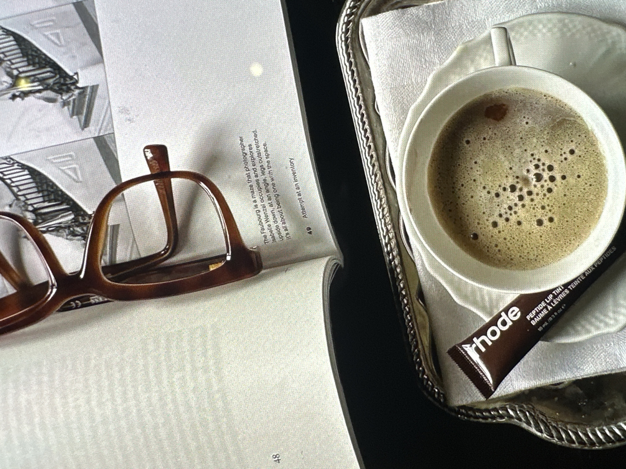 The image is an overhead shot of a book, eyeglasses, a cup of coffee on a silver tray with a white linen napkin, and a lip balm tube. The book is open to pages 48 and 49, with text visible and an architectural illustration. Brown-framed eyeglasses rest on page 48. The coffee cup is on a saucer on the tray, which also holds a dark brown lip balm tube labeled "rhode PEPTIDE LIP TINT." The background is a dark surface, likely a table. No people are visible. The location is not specified in the image.