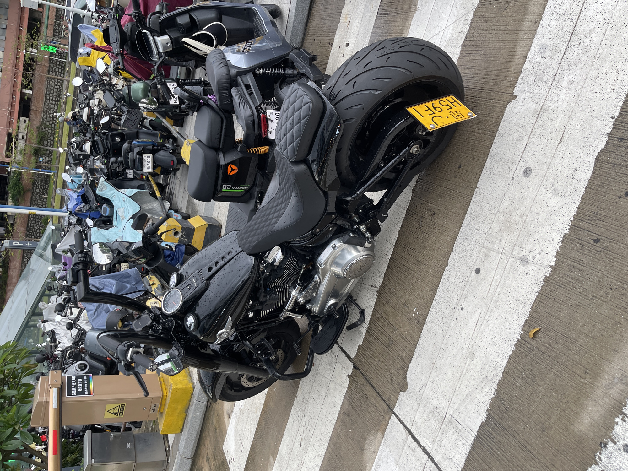 This media file shows a collection of parked motorcycles and scooters on a street, likely in Panyu District, China. The scene is an outdoor setting, with a paved surface marked with white lines, suggesting a designated parking area or a crosswalk. The time of day appears to be overcast, with the lighting suggesting it's either early morning or late afternoon. The weather seems to be damp, as indicated by water droplets visible on the motorcycles.
The main subjects are various types of motorcycles and scooters, predominantly black, with a prominent custom motorcycle in the foreground featuring a large rear tire and a yellow license plate. Several other smaller scooters and motorcycles are parked behind it. The surrounding environment includes a building with a stone facade and some greenery, possibly trees or bushes. There is also a parking barrier and an electronic kiosk visible, along with signage.
Visible text on the signage includes: "仅限商户送货车辆 及应急车辆" which translates to "Only for merchant delivery vehicles and emergency vehicles." Below this, there's a service phone number: "服务:020-31038666". Other visible text on license plates includes "HM6998", "61827", and "H59F1". There's also "02" visible on a part of one of the scooters, and "YEO" visible on a logo.
The overall impression is of a typical urban scene with a focus on parked vehicles in a functional area. There are no people directly visible in the main frame of the image. The image is captured from a slightly elevated, tilted perspective.