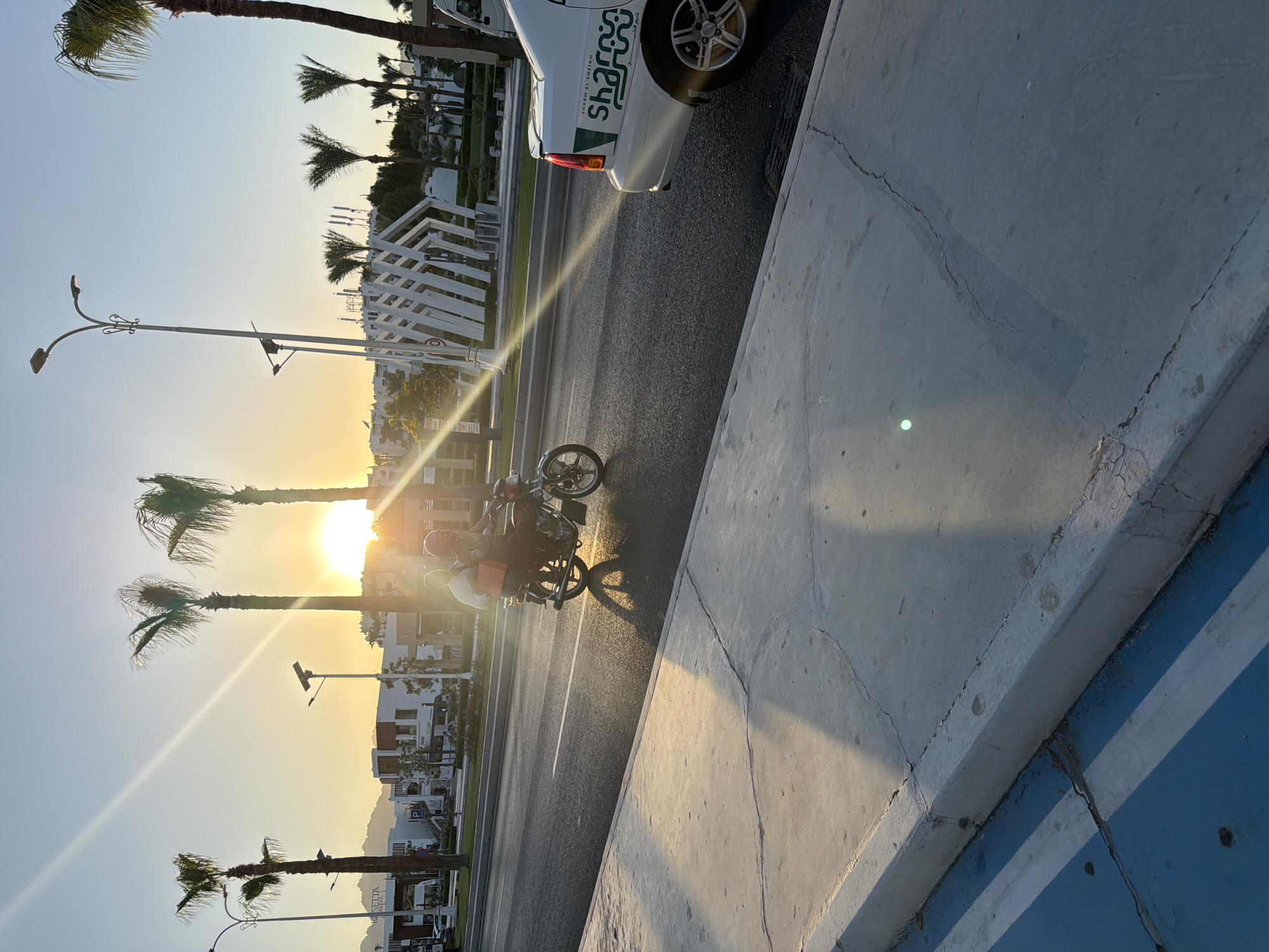 The image captures a street scene in Sharm El Sheikh, Egypt, during sunset. The sun is low on the horizon, casting long shadows and lens flare across the asphalt road. Two people are riding a motorcycle, with the passenger in the back holding an orange bag. A white car with "Sharm El Sheikh" written on its side is parked on the right side of the road. Palm trees and modern buildings line the street, with mountains visible in the background. The sidewalk in the foreground is made of concrete and shows signs of wear and tear, with some blue painted markings.