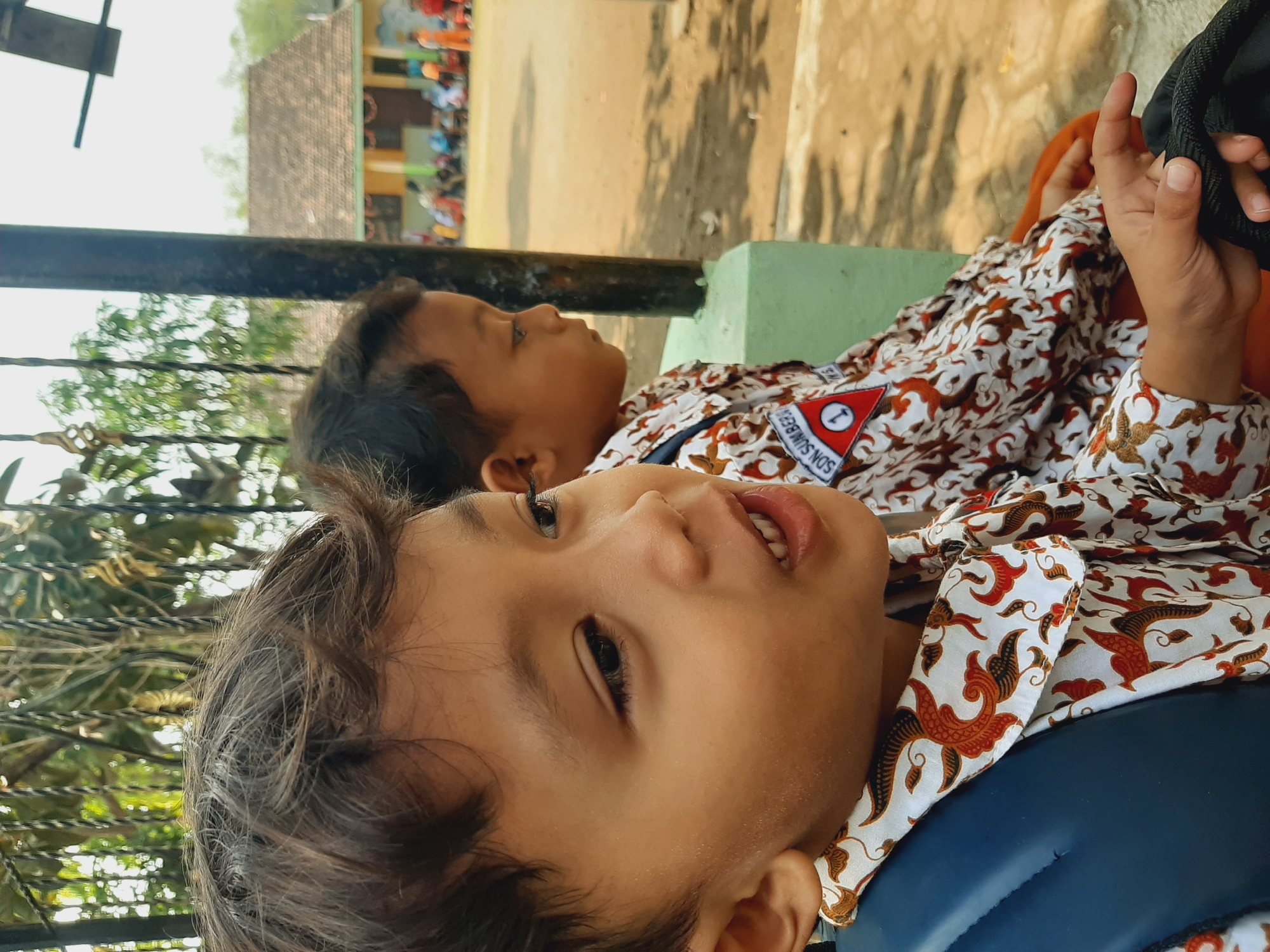 In a close-up, outdoor shot, two young boys are seated in the foreground, their faces turned to the right. The boy in the immediate foreground, on the left, has dark, tousled hair and is wearing a white shirt with a brown and red batik pattern and a blue backpack strap visible. His mouth is slightly open, and his eyes are looking to the side. He has a patch on his shirt that reads "SON. SUMBERJO" with a red triangle containing the number "1." The boy behind him, partially obscured, also wears a similar patterned shirt.

The background shows a school environment. A dark vertical post stands to the right of the boys, and beyond it, a building with a tiled roof is visible. Several other children and adults are blurred in the background, appearing to be gathered on a dirt or sandy area, suggesting a schoolyard or outdoor gathering space in Sumberjo, Indonesia. The overall lighting indicates it is daytime, with sunlight filtering through trees, casting dappled shadows.
