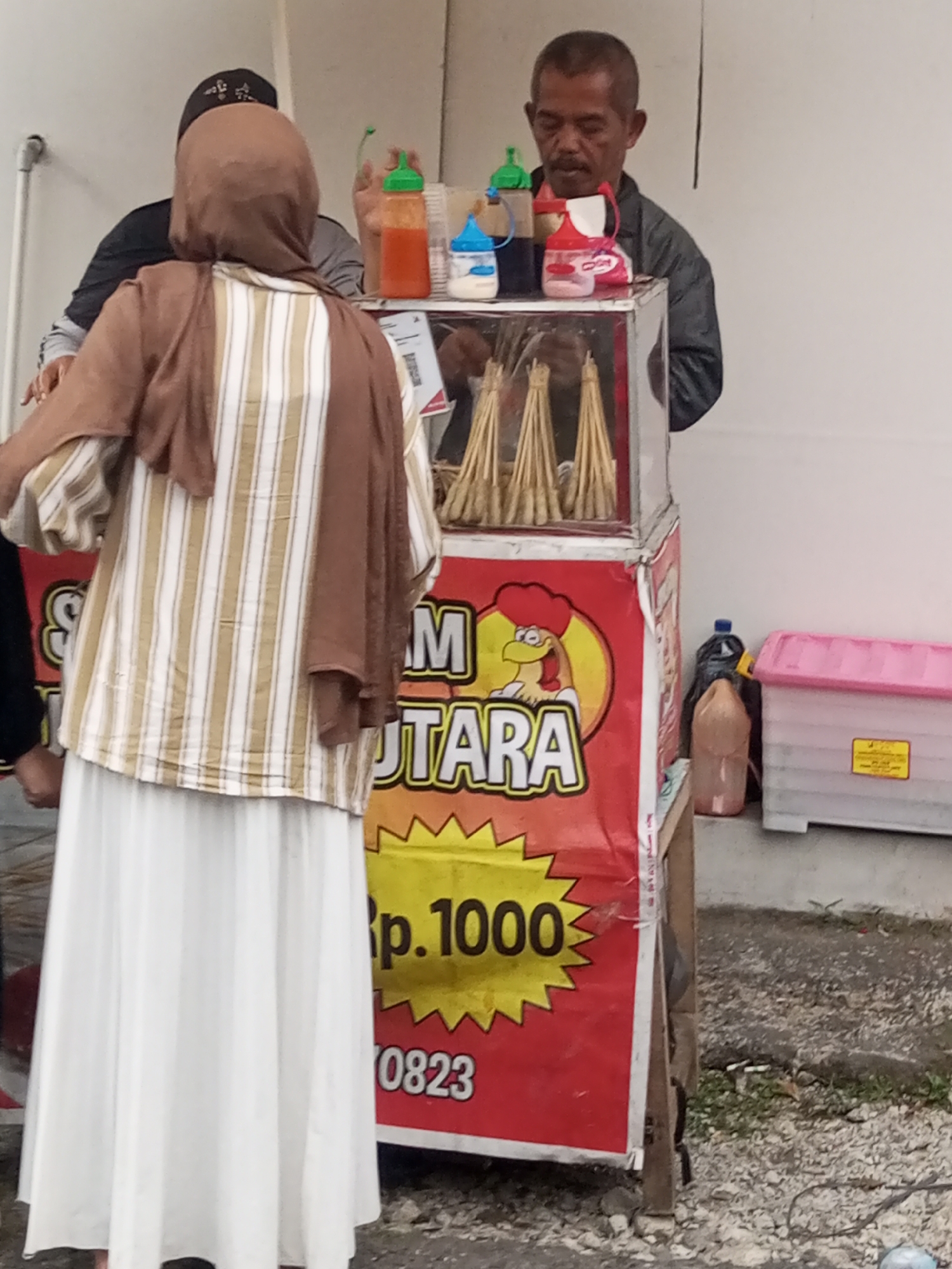 In Pandansari, Indonesia, a street vendor with dark hair and a mustache is operating a food cart. The cart is adorned with a red banner featuring a cartoon chicken and text that reads "SAM UTARA RP. 1000 0823". On top of the cart are several bottles of condiments, including red, blue, and clear liquids. A woman wearing a brown hijab and a striped shirt stands in front of the cart, facing away from the camera. Her attire includes a long, white skirt. Behind the woman, another person is partially visible, wearing a black head covering. To the right of the food cart, a pink and white storage container is present, along with a dark plastic bag and a bottle. The ground is covered in gravel and dirt.