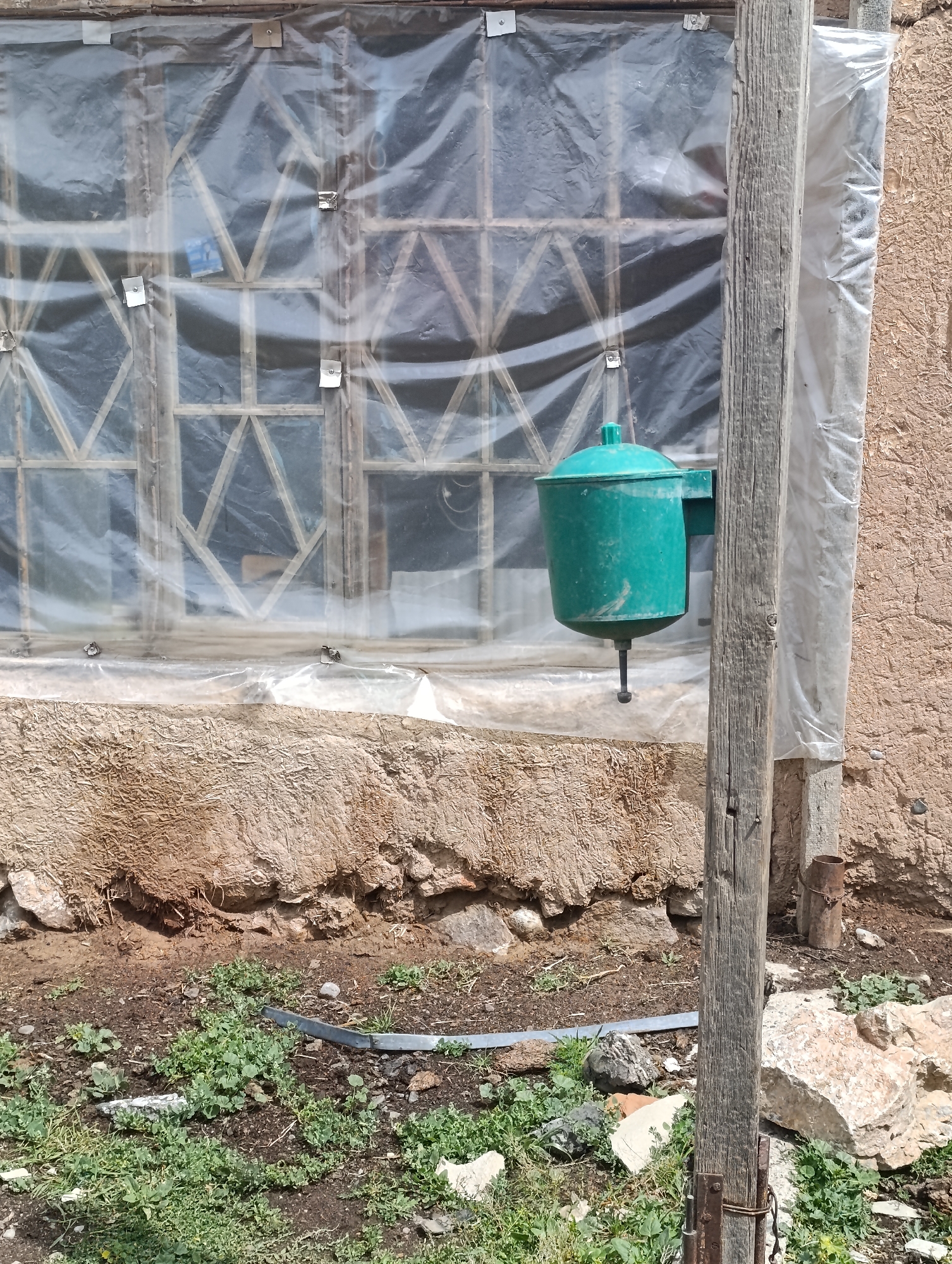 The image shows a rustic building in Ак Жар, Kyrgyzstan. The building appears to be constructed with earth and straw, and has a weathered appearance. A large window is covered with clear plastic sheeting, which is secured with metal clips. A green plastic watering device with a spout is attached to a wooden post to the right of the window. The ground in front of the building is covered with dirt, small green plants, and rocks. A metal pipe runs horizontally across the ground. The overall scene suggests a rural or agricultural setting.
