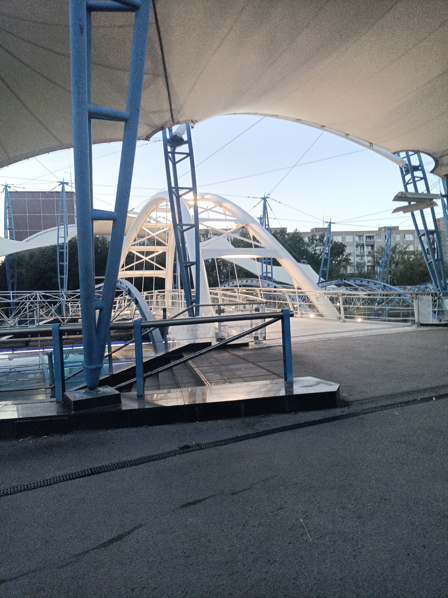 The image depicts an exterior scene under a large, light-colored canopy structure supported by blue metal beams. The canopy is taut and stretched, providing shade. In the background, a white, arched bridge with intricate metal supports is visible, illuminated by lights. The bridge appears to be part of an elevated walkway or structure. Surrounding the bridge and canopy are trees and apartment buildings, indicating an urban setting. The ground in the foreground is paved with asphalt, with a faint dark chain-like pattern embedded in it. The lighting suggests it is either early morning or late evening, with a clear, pale blue sky. The scene appears to be a public space, possibly a park or an outdoor recreational area, characterized by modern architectural elements. The image is taken at a slightly low angle, looking up towards the canopy and bridge. There are no people visible in the image.