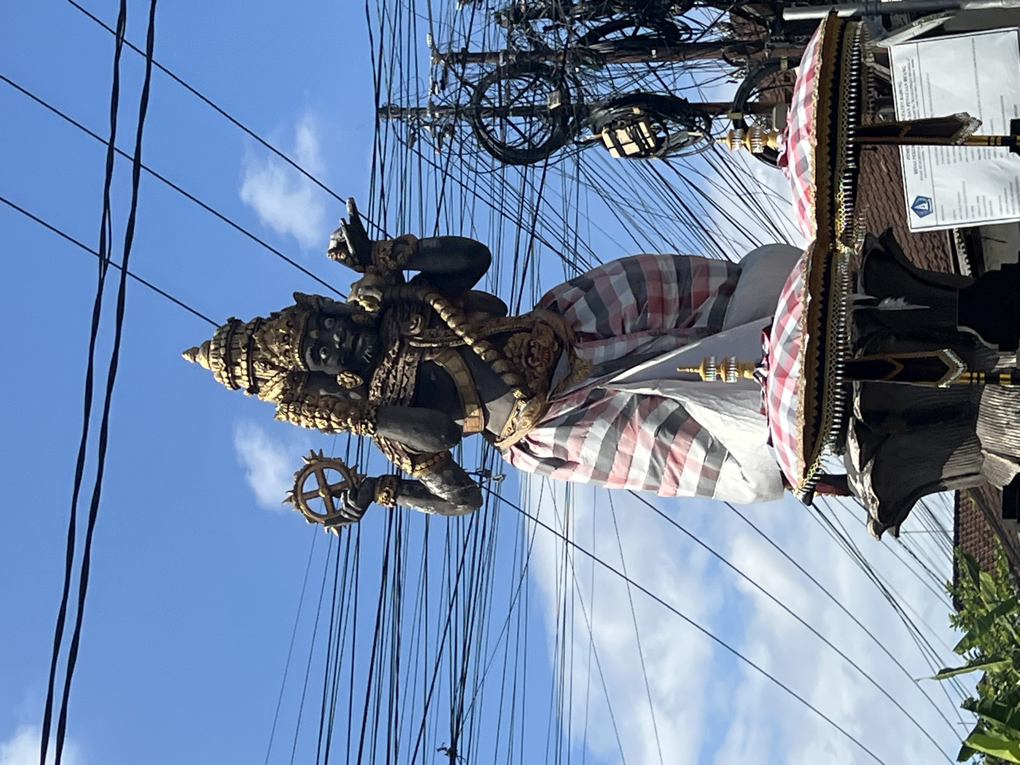 A large, dark-skinned statue adorned with gold accents and a striped sarong stands against a bright blue sky filled with a tangled web of electrical wires. The statue holds a golden wheel in its left hand and appears to be wearing a golden headdress and jewelry. The lower portion of the statue is decorated with two ornamental umbrellas with checkered patterns. In the background, there is a utility pole heavily laden with electrical cables, and some greenery is visible at the bottom left. The overall scene suggests an outdoor setting, likely a street in North Kuta, Indonesia, characterized by its vibrant cultural statues and the pervasive presence of overhead wiring.