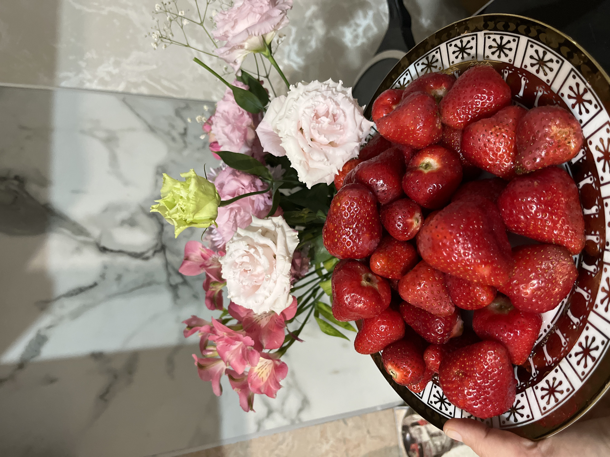 The image displays an indoor scene focused on a plate of fresh strawberries held by a human hand, set against a background of a floral arrangement and a tiled wall.
In the foreground, a round, shallow ceramic plate is filled with numerous ripe, red strawberries. Many of the strawberries exhibit a glistening surface, indicating moisture, and some retain small green caps. The plate has a distinctive decorative rim featuring a repeating pattern of white square sections containing a dark, stylized star or snowflake motif, all bordered by a metallic gold band. The center of the plate is a solid dark reddish-brown color. A fair-skinned hand is visible on the lower-left side, holding the plate by its rim.
Behind the plate, a bouquet of fresh flowers is arranged. The bouquet primarily features light pink and white Lisianthus flowers with ruffled petals, several smaller pink Alstroemeria blooms, and a single pale green/yellow Lisianthus bud or flower. Delicate white Gypsophila (Baby's Breath) is also interspersed throughout the arrangement.
The background consists of light-colored, vertically oriented wall tiles with gray marble-like veining, consistent with a kitchen or bathroom backsplash. A prominent vertical grout line is visible towards the center of the background. Reflections on the tiles suggest an overhead light source. On the far right, a dark, blurred object, possibly an appliance or countertop edge, is partially visible. On the far left, a faint, blurred metallic object is discernible.
There are no discernible details within the image to confirm the location as the city of Kaspiysk, Russia.