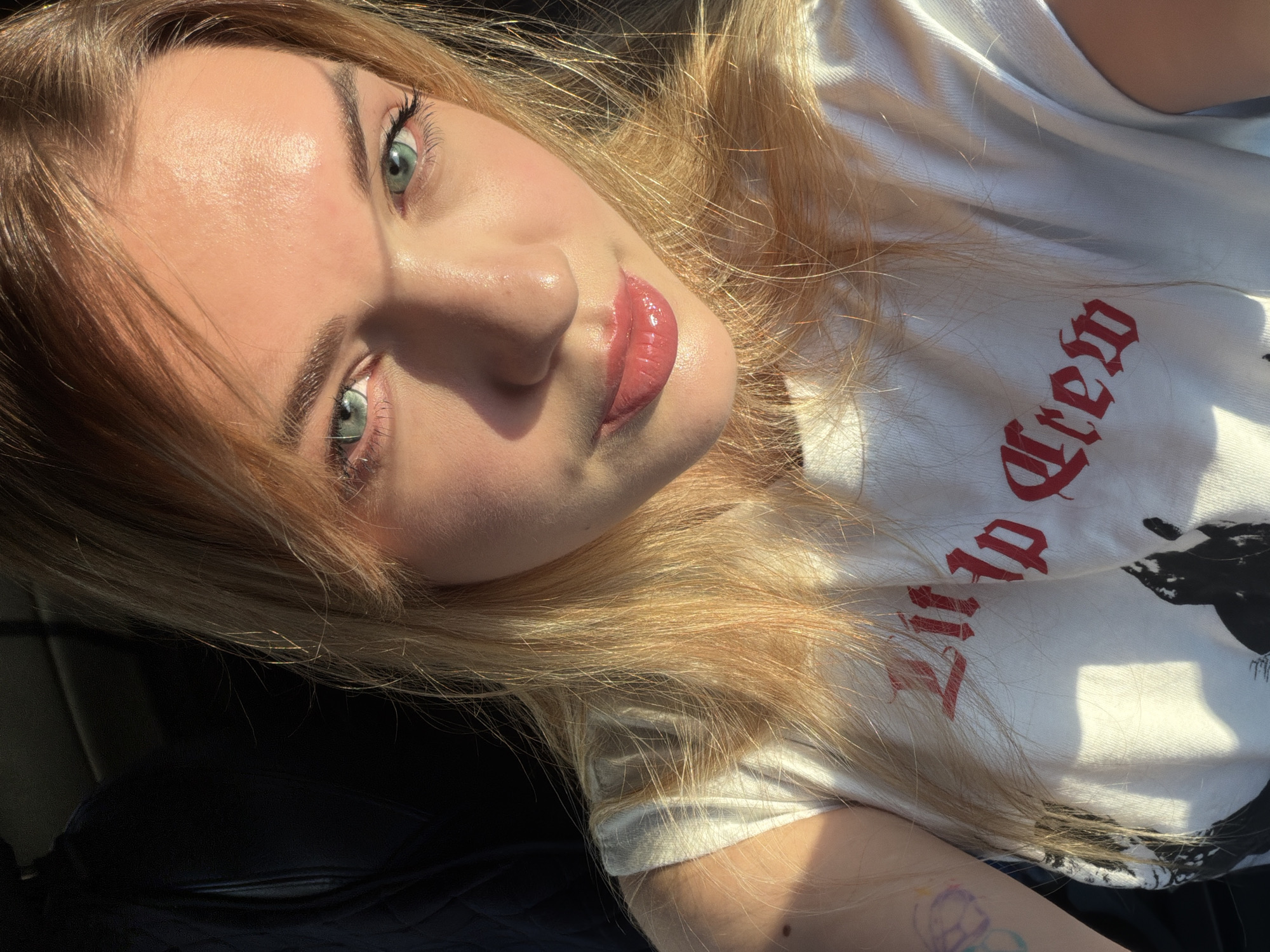 A young, light-skinned person with blonde hair and green eyes is lying on a black surface. The person is wearing a white t-shirt with red writing that says "Mad Crew". The lighting appears to be natural sunlight. A tattoo is visible on the person's left arm. The location of the image is unknown, and there is no indication that it was taken in Naberezhnye Chelny, Russia.