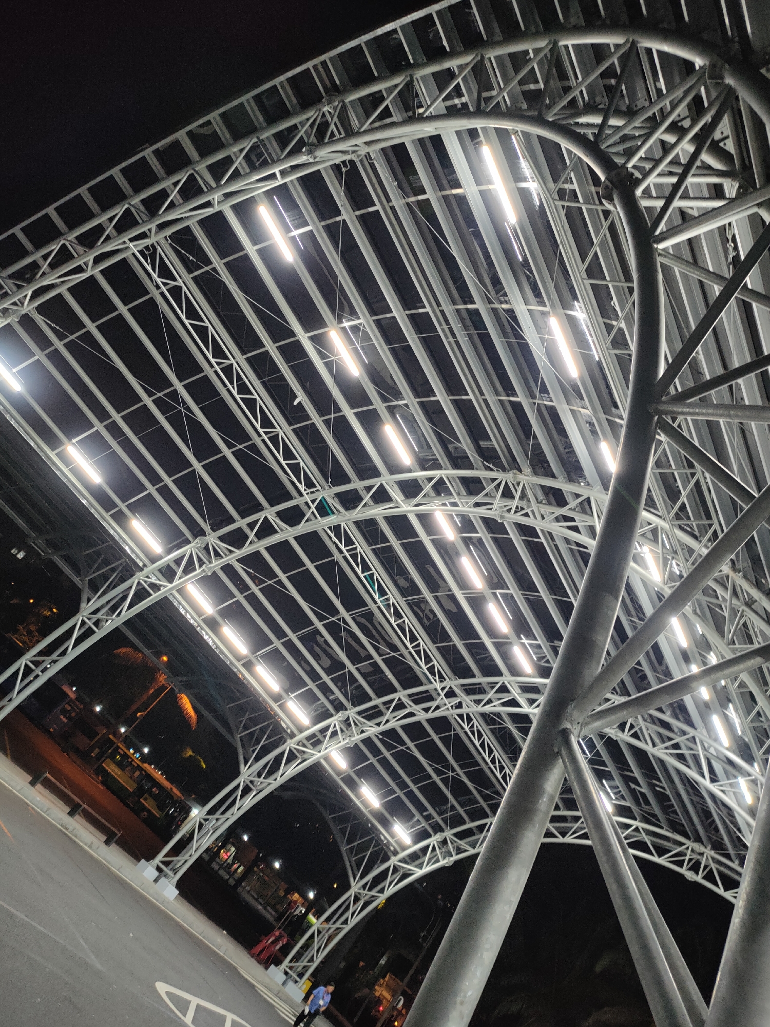 The image depicts a large, modern, covered walkway structure at night in Thủ Đức, Vietnam. The walkway's roof is supported by a complex steel truss system, illuminated by numerous bright, linear LED lights. The perspective is from below, looking upwards at the intricate metalwork and the rows of lights against a dark sky.
In the background, a street or parking area is visible. A yellow bus with visible text, possibly advertising, is parked on the left. Several palm trees with distinctive fronds are also present, suggesting a tropical or subtropical environment. The ground is paved, with white markings indicating a road or parking area. A single person, dressed in a blue shirt and dark pants, is visible on the paved area in the lower right corner, walking away from the camera. The overall scene suggests an outdoor public space, possibly a bus station, market, or transportation hub, that is operational and well-lit at night. The lighting creates a bright, almost futuristic atmosphere within the covered area, contrasting with the darkness of the night sky. The weather appears clear.