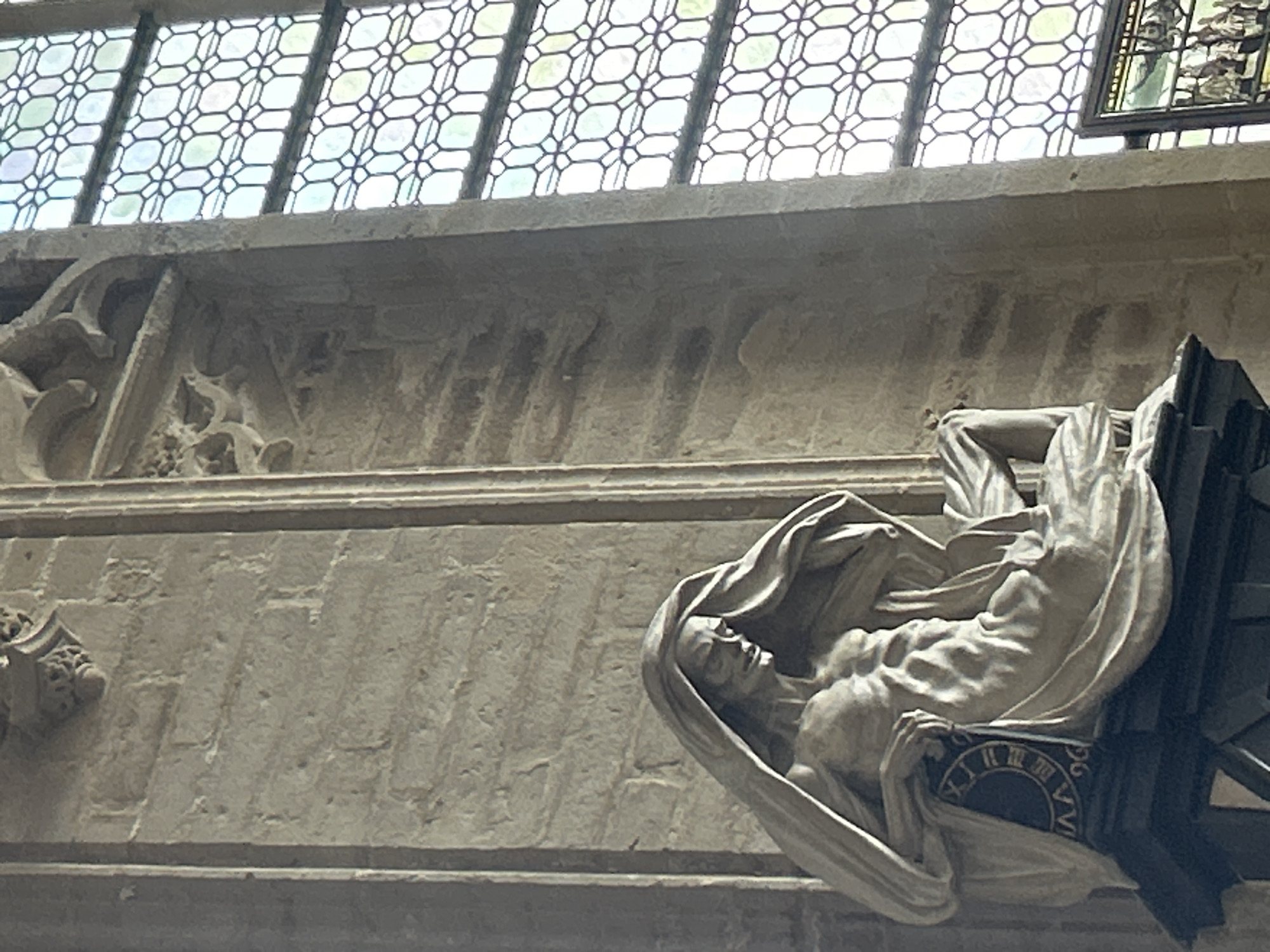 The image shows a marble sculpture of a gaunt, draped figure seated on a pedestal with a dark circular object featuring Roman numerals. The sculpture is located in the Saint Peter's Church in Leuven, Belgium. To the right of the sculpture is a large stained-glass window with a repeating geometric pattern of octagons and squares. The wall behind the sculpture is made of rough, light-colored stone blocks. In the bottom right corner, a portion of another stained-glass panel depicting a scene with multiple figures is visible. The lighting suggests it is daytime, with light streaming in from the window.