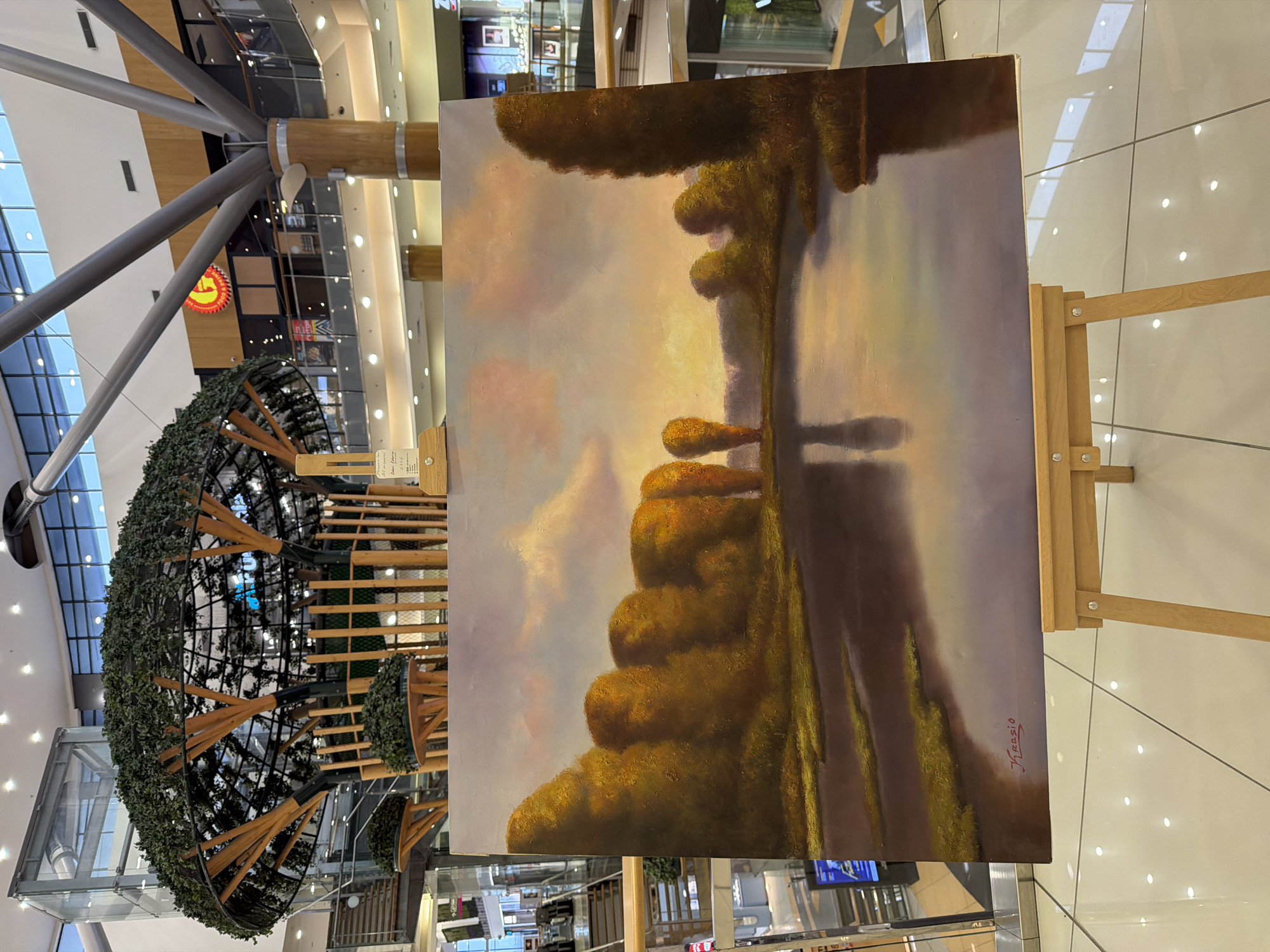An oil painting, signed "J. Krasio" in red on its lower-left corner, depicts a serene landscape at dusk or dawn with a body of water reflecting a pastel-colored sky and diffused clouds. The water is bordered by a continuous line of rounded trees or dense foliage in autumnal shades of yellow and orange. The painting rests on a light brown wooden tripod easel. This display is situated on a glossy, light-colored tiled floor within a brightly lit, multi-story indoor commercial complex, identified as a shopping mall in Unknown, Unknown.
In the background, a large, multi-level decorative structure resembling a tree, constructed with a spherical metal cage and covered in artificial dark green foliage, is prominent. Large, silver-colored radial support beams extend from a central column towards the ceiling. Upper levels of the mall are visible, featuring glass railings and various storefronts, including one with a circular red and yellow logo containing the letter "G". A glass elevator shaft is visible on the left. A small, partially obscured sign attached to the easel behind the painting displays text that includes "Original" and "2000." No people are visible within the frame.