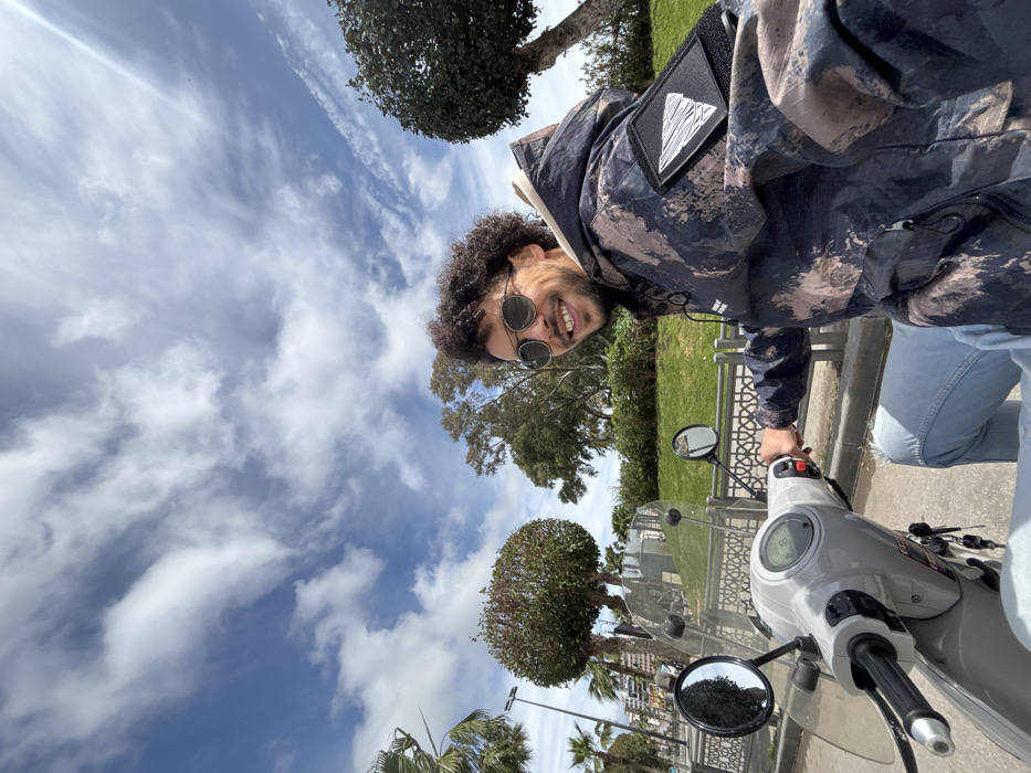 A male individual is seated on a light grey scooter under a blue sky with scattered white clouds. He possesses dark curly hair, a short beard, and wears round sunglasses, a multi-patterned hooded jacket with a white triangular patch on the left sleeve, and light blue ripped jeans. The man is smiling and looking towards the camera, with his right hand positioned on the scooter's handlebar. The scooter features a clear windscreen, visible rearview mirrors, and a digital dashboard, with keys in the ignition. The background includes a grassy area with various trees, including neatly trimmed spherical bushes, bordered by a decorative metal fence with geometric patterns. In the distance to the left, palm trees, streetlights, and buildings are visible. The scene is located in a park or promenade area within the city of Tanger, Morocco.