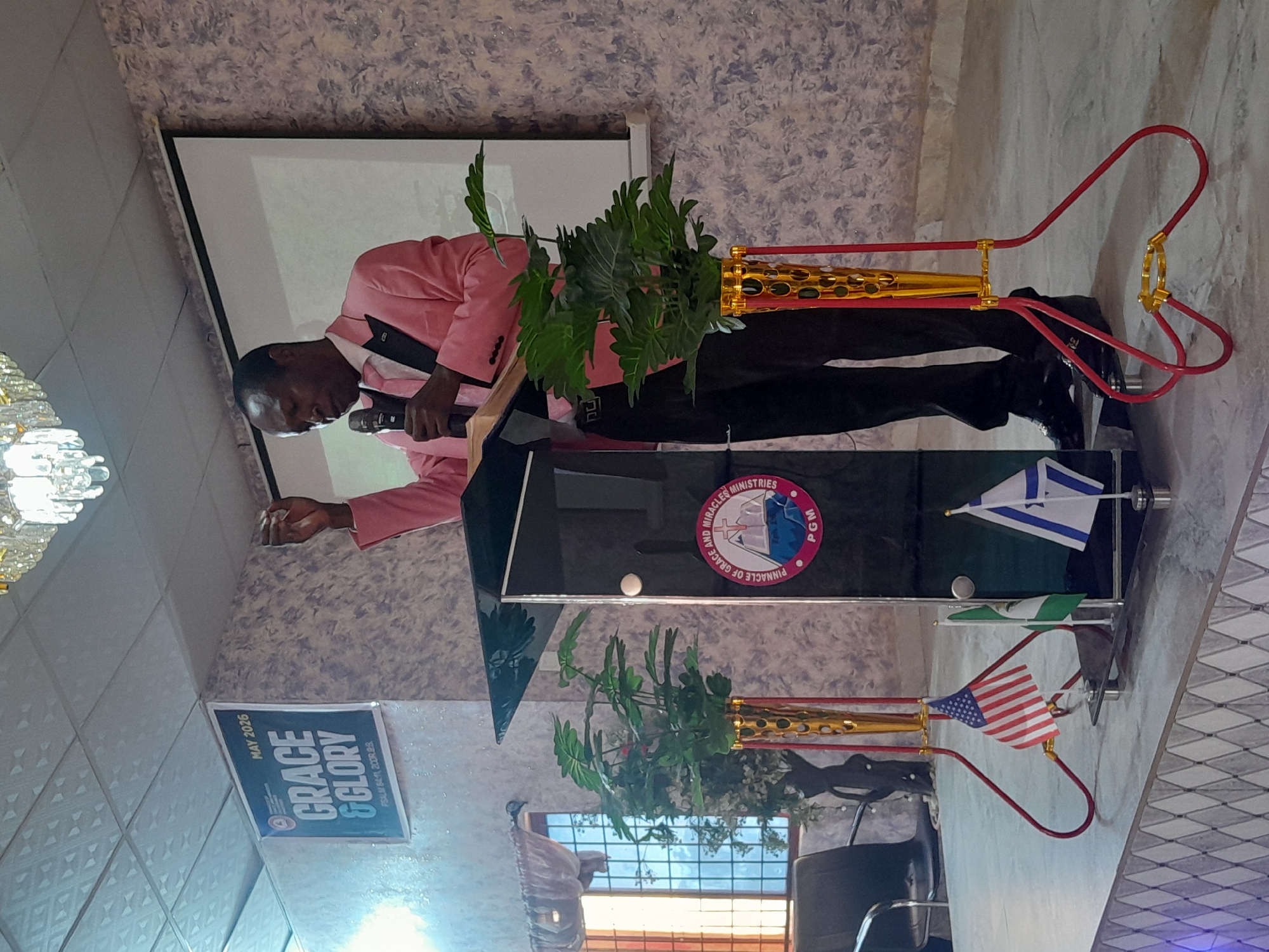 The image captures a man, likely a pastor, standing behind a black lectern, presumably delivering a sermon. He is dressed in a pink suit jacket over a black shirt and pants, and he holds a microphone in his left hand. The lectern is adorned with the emblem of "Pinnacle of Grace and Miracles Ministries (PGM)," miniature flags of the United States, Nigeria, and Israel. The background features wallpaper and a projector screen. A sign indicating "Grace and Glory, MAY 2026," referencing Psalm 84:11, is posted on a nearby wall. This is located in the city of Jos, Nigeria.