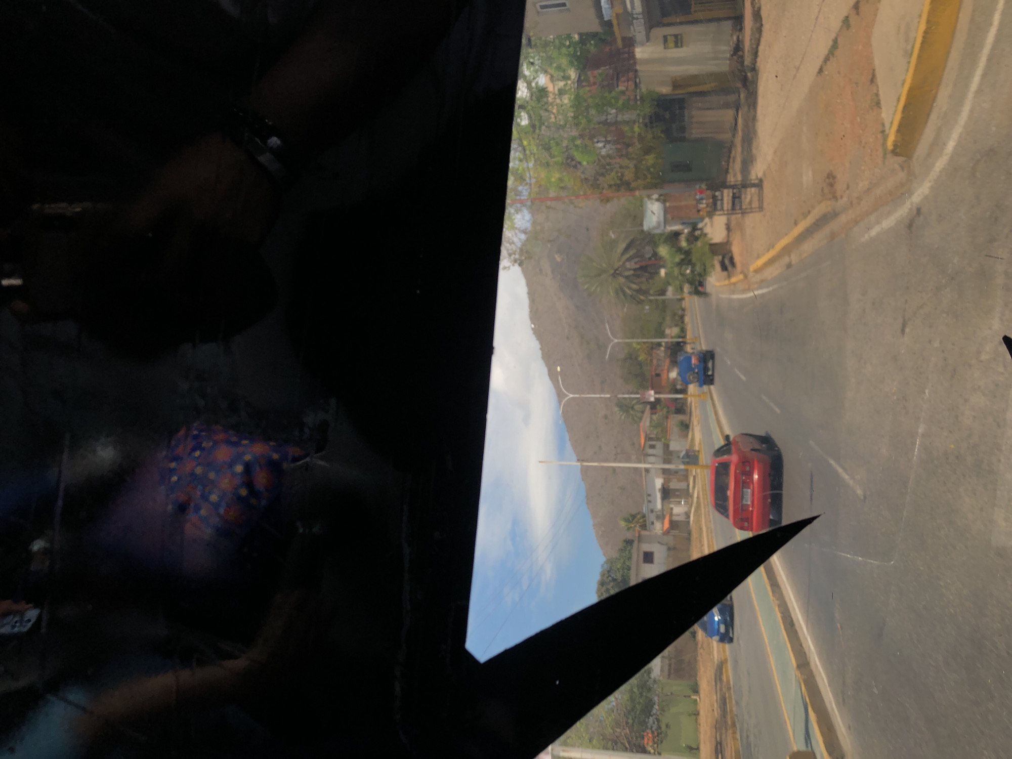 The photograph shows a view from inside a vehicle, looking out onto a street scene in El Valle del Espíritu Santo, Venezuela. The viewer is likely inside a vehicle (bus/car), the road runs alongside a mountain. Cars are visible on the road. Part of the vehicle's interior (arm, window frame, and headrest) is visible in the frame. The presence of buildings and other structures suggests a populated area. The sky is visible.