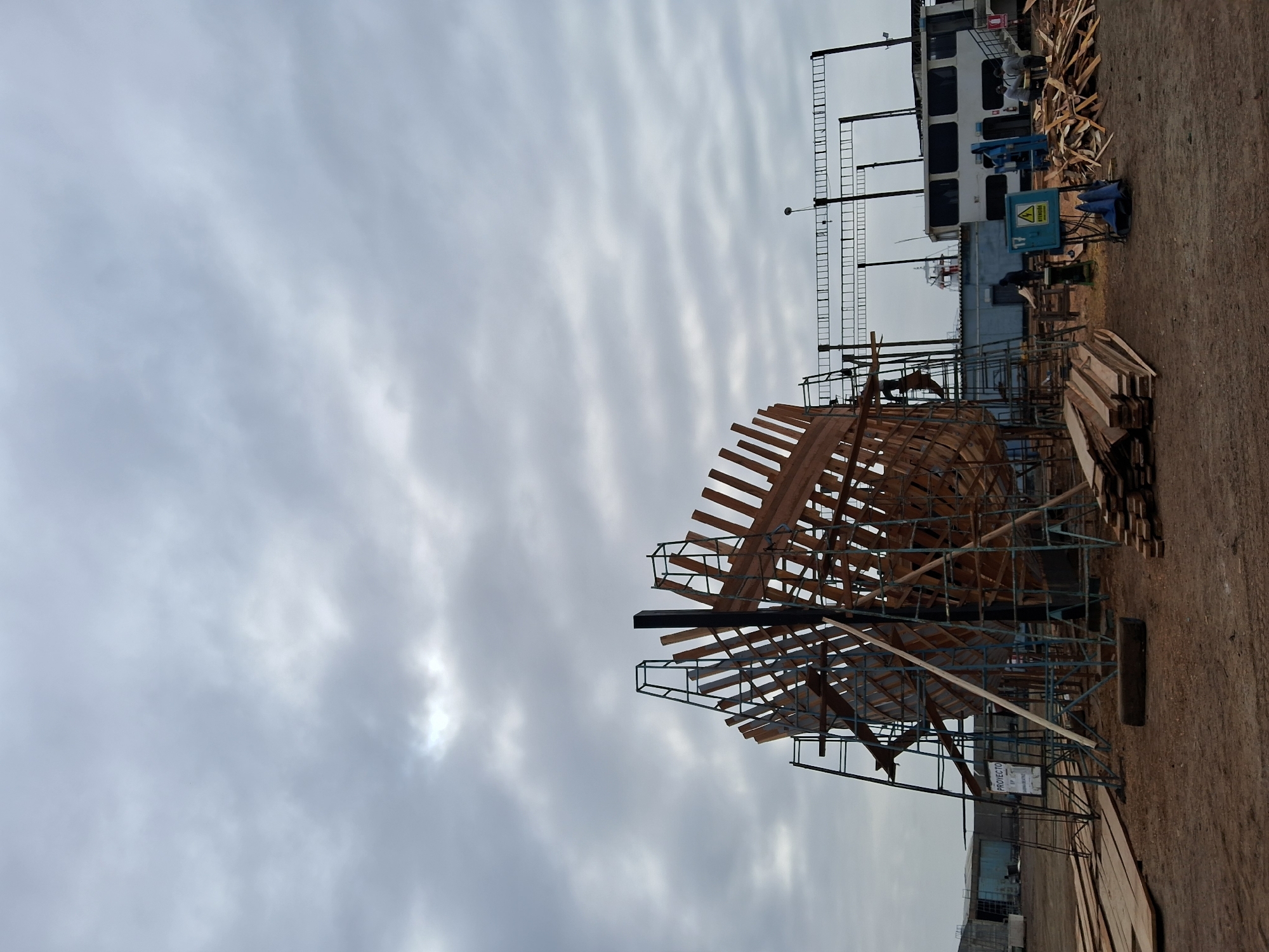 In Chimbote, Peru, a shipyard is captured under an overcast sky. The central focus is the wooden hull of a boat under construction, supported by scaffolding. Piles of lumber are scattered across the dusty ground, with planks forming a makeshift walkway near the hull. In the background, industrial buildings and a ferry or boat are visible. Several individuals are present, some working on the construction, while others are standing around in conversation. A sign with "PROYECTO" is displayed on the left, indicating the project. The overall scene depicts a typical day at a bustling maritime construction site.