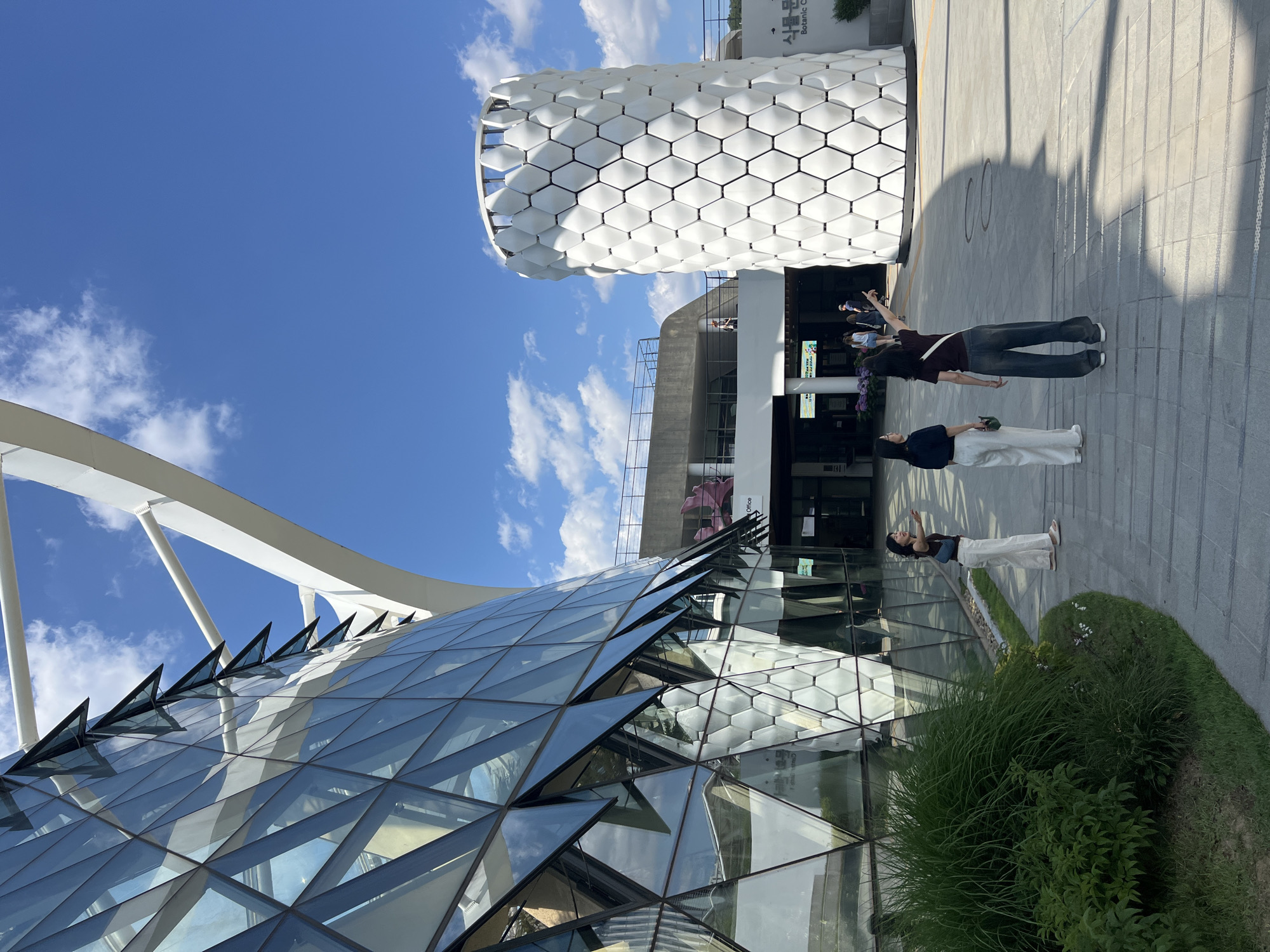 The image captures a sunny, clear day at a modern architectural complex. The scene features two prominent buildings, three individuals, and paved walkways amidst some greenery.
On the left, a large, contemporary building features an intricate glass-paneled facade, appearing as a geodesic or faceted dome structure. Its surface is composed of numerous triangular glass sections that reflect the bright blue sky and scattered white clouds. White, curving structural elements arch gracefully over and around this glass edifice.
To the right, a cylindrical or partially cylindrical building stands tall, distinguished by its striking white exterior. This facade is covered in a uniform pattern of raised hexagonal elements, creating a three-dimensional, honeycomb-like texture. Visible text on this building reads "식물문화" (Plant Culture) and "Botanic Cafe," suggesting its function.
On the paved area in the foreground and middle ground, three individuals are present. They are casually dressed and appear to be observing the architecture, with their gazes directed upwards towards the buildings. One person, wearing a dark top and dark pants, has their arms slightly raised, seemingly gesturing or taking a photo. Another person in a dark top and light pants stands nearby, also looking up. The third individual, further left, is similarly engaged.
The time of day is bright daylight, evidenced by the clear blue sky, scattered white cumulus clouds, and strong shadows cast by the buildings and people on the paved ground. Lush green bushes and grass are visible in the foreground and along the base of the buildings, adding a natural element to the structured environment. The overall impression is one of modern design and an open, inviting public space.
