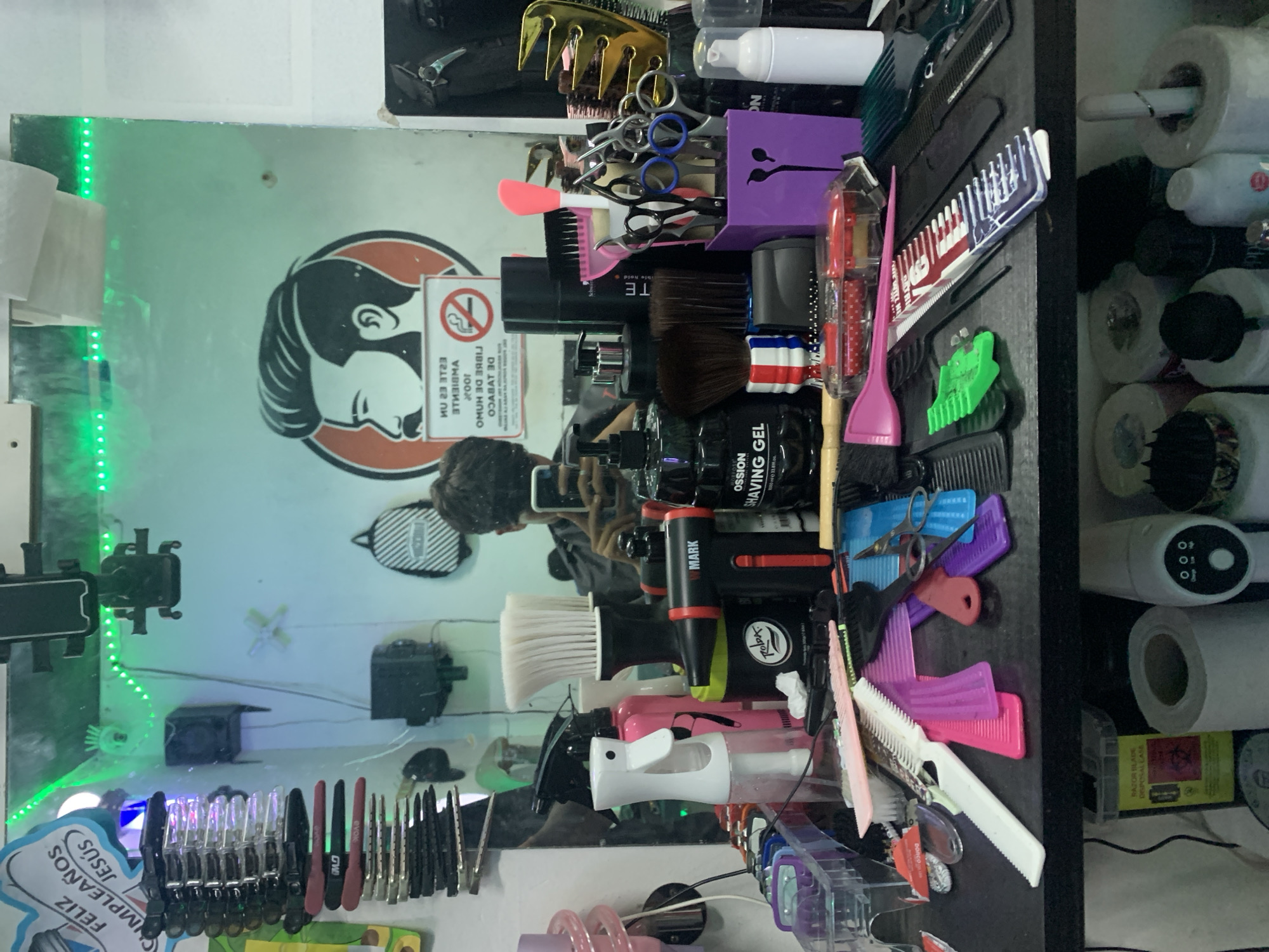 This image captures a close-up view of a barbershop counter and its reflective mirror, situated in Tacarigua, Venezuela. The counter is densely populated with a variety of professional grooming tools and products.
In the immediate foreground on the black counter, numerous combs are haphazardly arranged, varying in color (pink, purple, white, black, green) and design. Several pairs of scissors are visible, with some stored upright in a purple container on the right side. Various brushes are present, including neck dusters, a shaving brush with a red, white, and blue barbershop pole-striped handle, and other hair brushes. Multiple spray bottles, one white and one pink, are positioned on the left side of the counter. Hair clippers and their attachments are organized in a clear plastic container. Product bottles include a black pump dispenser bottle labeled "OSSION SHAVING GEL" and a black tub labeled "Rolpa." A black and red handheld device, identified as "MARK," lies near the center. A pink spatulate tool and a small clear container with red accessories are also visible.
The large mirror in the background reflects the upper portion of the barbershop environment. A reflection of an individual, presumably the photographer, is visible in the lower-right quadrant of the mirror, holding a smartphone. Prominently reflected on the mirror's surface is a circular graphic depicting a stylized profile of a man with a beard and swept-back hair. Below this graphic, a "NO SMOKING" sign is reflected, displaying the Spanish text "ESTE ES UN AMBIENTE 100% LIBRE DE HUMO DE TABACO" (This is a 100% tobacco smoke-free environment). Green LED strip lights run horizontally along the top edge of the mirror. Mounted above the mirror are several black smartphone holders. To the far left, a white sign with blue lettering, "FELIZ CUMPLEAÑOS JESÚS" (Happy Birthday Jesus), is partially visible. A small black and white striped backpack hangs on the wall, reflected in the mirror. Small black speakers are also mounted on the wall and reflected.
Below the main counter, a lower shelf contains additional items. These include multiple rolls of paper, a yellow plastic bin labeled "RAZOR BLADE DISPOSAL" with a biohazard symbol, and various bottles and containers. A white electronic device with a black control panel and indicator lights is also present.