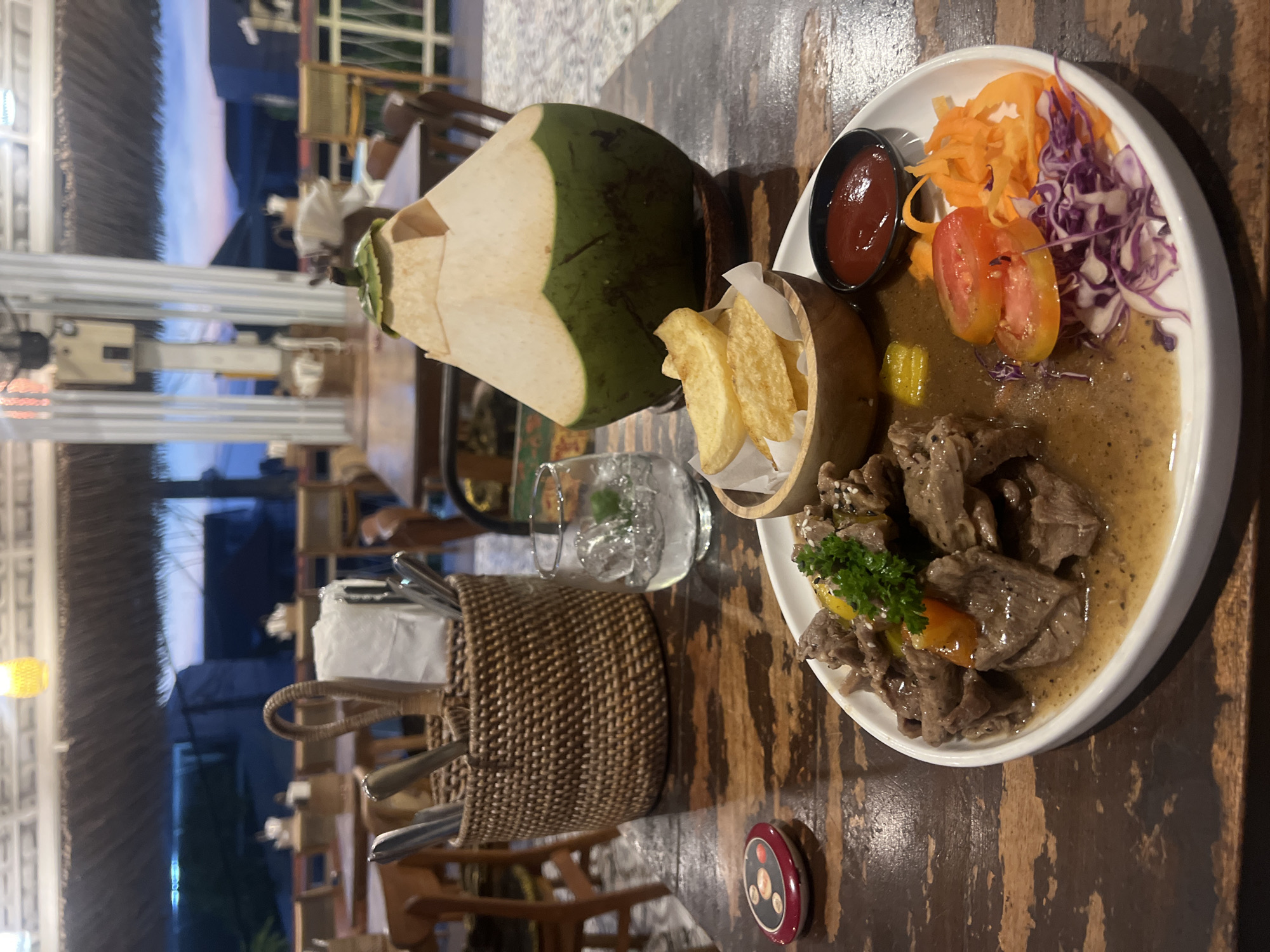 This image captures a close-up of a meal served at a restaurant in North Kuta, Indonesia, during the evening. The scene is set on a dark, rustic wooden table, suggesting an informal or tropical dining atmosphere.
The central focus is a white oval plate featuring a generous portion of cubed meat, likely beef, served in a light brown, possibly peppery, sauce. The meat is garnished with fresh green parsley, small pieces of yellow bell pepper, and sprinkled with white sesame seeds. On the same plate, a side salad provides a colorful contrast with shredded orange carrots, thinly sliced red tomatoes, and shredded purple cabbage. A small black bowl containing red sauce, presumably ketchup, is also present on the plate.
Adjacent to the main course, a small, light-colored wooden bowl holds a serving of golden-brown french fries. To the left of the food, a large, fresh green coconut, partially cut to reveal its white interior, stands upright with a straw, indicating a natural beverage. Beside it, a clear glass filled with ice and a slice of lime suggests a refreshing drink, possibly water. A woven rattan basket, filled with silverware including forks and spoons, sits between the drinks and the main dish.
The background, largely out of focus, reveals elements of the restaurant's structure, including what appears to be a thatched roof. The sky, visible in the distance, displays a purplish-blue tint, characteristic of dusk or early evening. Artificial lights are also discernible in the background, further confirming the time of day. A small, circular red and black object with white symbols, possibly a coaster or a service call button, is visible on the table in the foreground. The overall impression is that of a relaxed and enjoyable dining experience in a tropical setting.