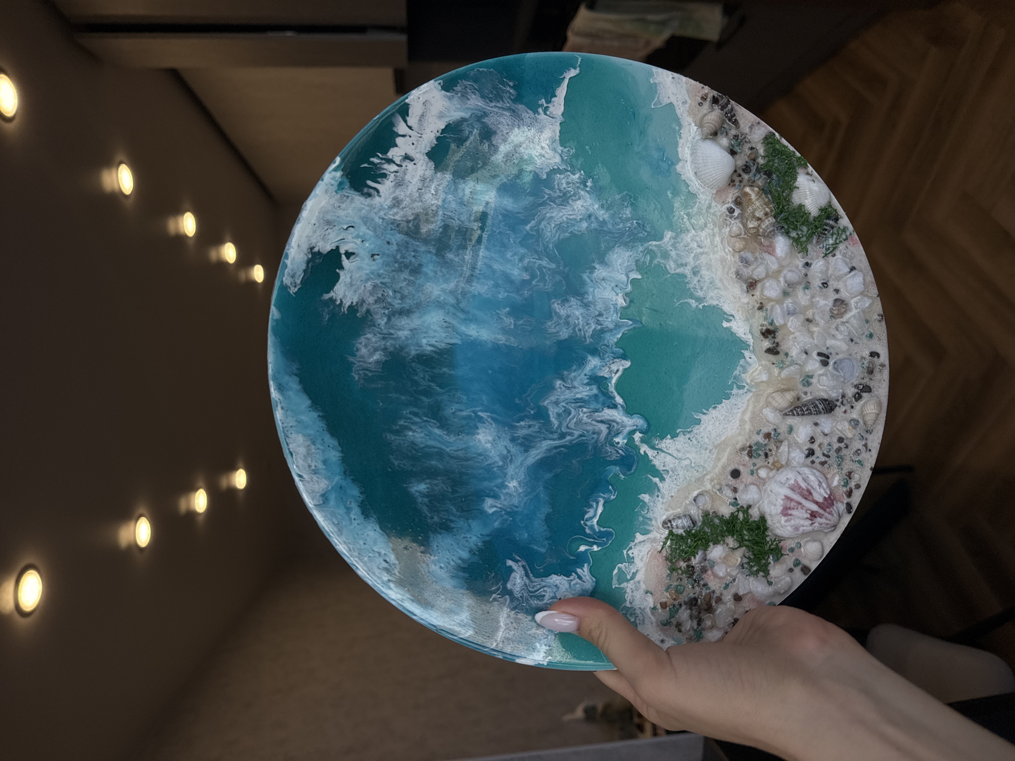 A hand holds a round art piece resembling an aerial view of a coastline. The artwork is made of resin and features a blue and white "ocean" area with a sandy "beach" area adorned with small pebbles, seashells, and patches of green vegetation. The hand has manicured nails with a white French tip. The background is a dimly lit room with a row of recessed lights on the wall. The location is unknown.