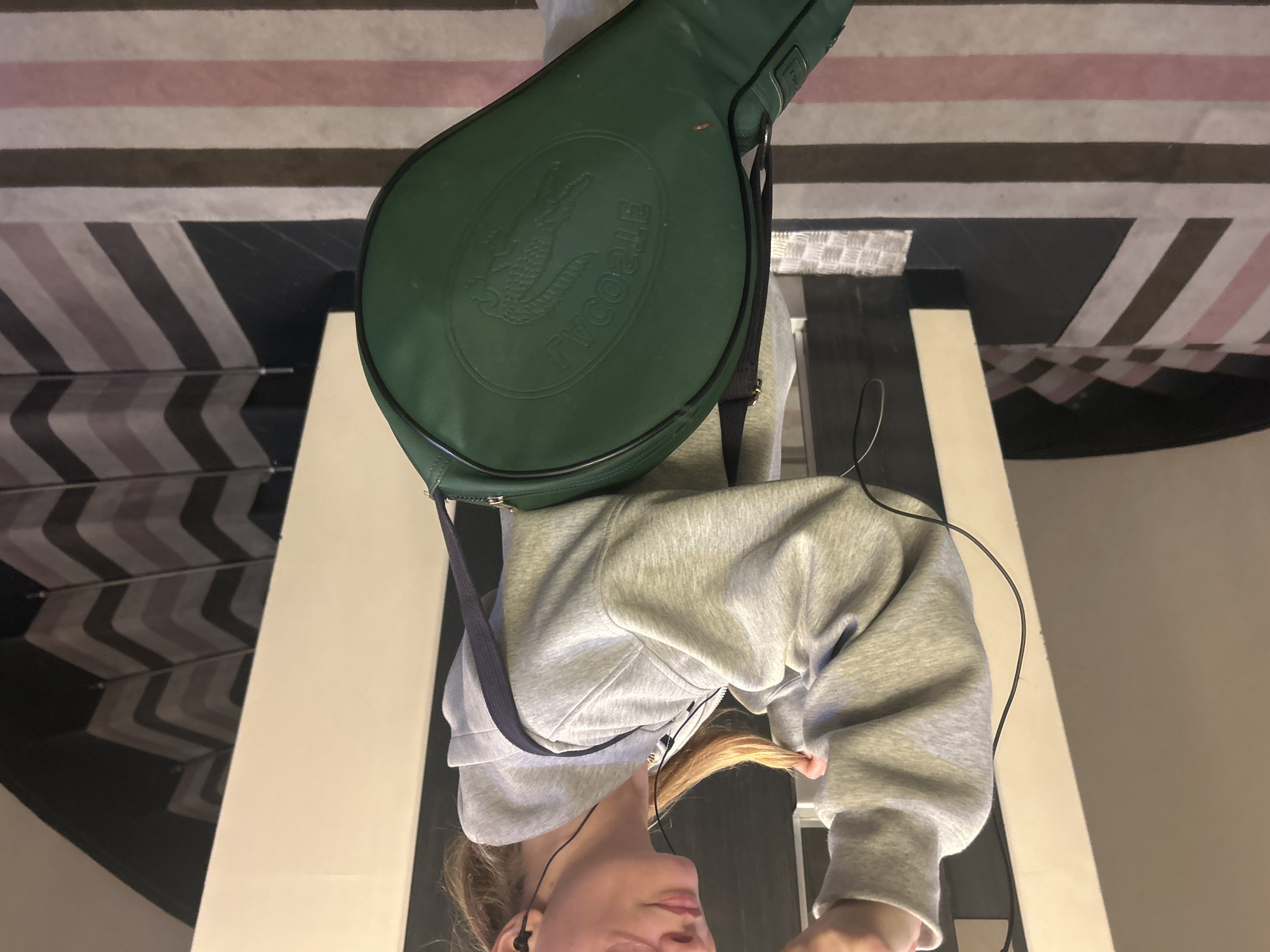 A person is carrying a green Lacoste tennis racket bag. The person is wearing a grey hoodie with a blue strap and headphones. The location is an interior space featuring a wall or ceiling with striped patterns.