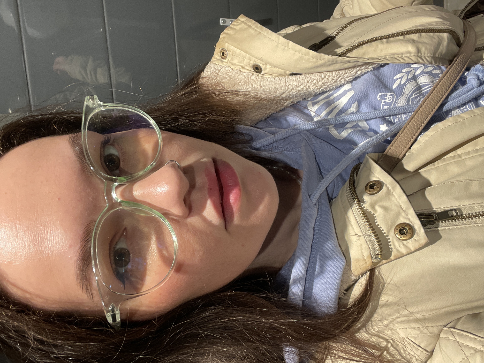 The image, rotated 90 degrees clockwise, presents a close-up portrait of a young adult female. She has long, dark brown hair and is looking directly at the camera through clear-framed glasses. Her eyes are light-colored, and she wears a small silver nose ring on her left nostril. Her lips are a natural pinkish-red, and her skin appears clear with a subtle sheen on her forehead and cheeks, suggesting strong lighting.
She is dressed in a light beige or khaki jacket with a visible fuzzy, possibly sherpa-lined, collar and metallic snap buttons. Underneath, a light blue hooded sweatshirt is visible, featuring white graphic prints that resemble a crest or emblem with some illegible text and symbols. Two light blue drawstrings hang from the hoodie. A beige, textured leather-like strap, likely from a purse, crosses over her jacket. The jacket also has prominent zippers and stitching, and a small discolored or stained area is noticeable near a zipper closure on the right side.
The background is a plain grey tiled wall, which reflects some light. The lighting is strong and directional, illuminating the left side of her face and casting shadows on the right. A faint reflection, possibly of a hand or arm, is visible on the grey wall in the upper left area of the frame. The subject maintains a neutral expression. The scene is set indoors, in Izhevsk, Russia.