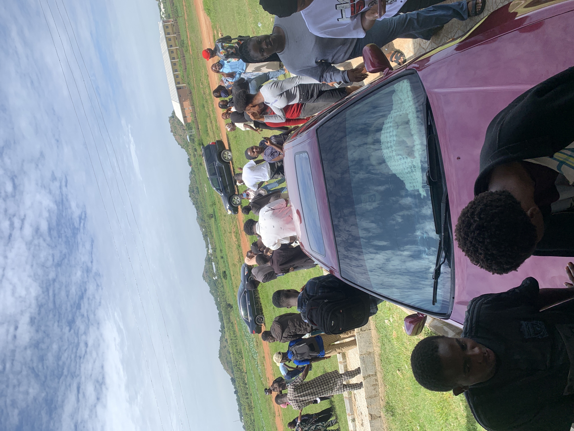 The image captures a group of people gathered outdoors in Gwafan, Nigeria. The scene appears to be informal, possibly a social gathering or a casual meeting. Several individuals are standing and sitting around a maroon-colored car, with some looking towards the camera. The ground is a mixture of grass and dirt, suggesting a rural or semi-rural setting. In the background, a large building with multiple windows and a light-colored roof is visible, potentially a school or an administrative building. The sky is partly cloudy, indicating daytime. The overall mood seems relaxed and communal, with people engaging with each other. There is no visible text in the image.