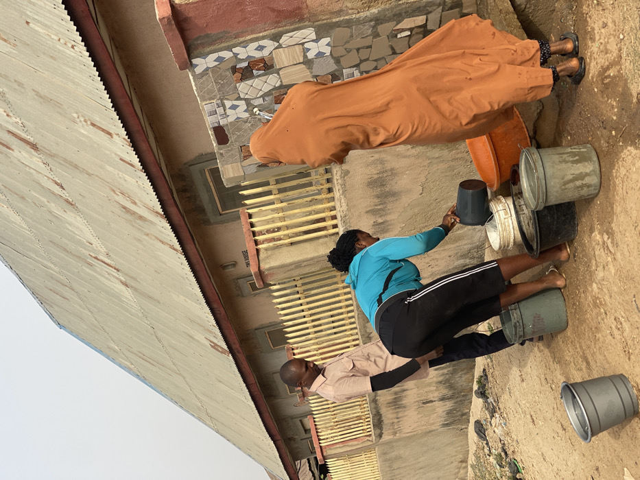 Three individuals are depicted alongside a house exterior. A woman in an orange robe stands by a bucket. Another woman, wearing a blue jacket and black pants, fills a dark-colored bucket. A man in a light-colored shirt stands nearby. Several other buckets are stacked and scattered around the ground, some filled with water. The house features a metal roof, tiled exterior details, and barred windows.