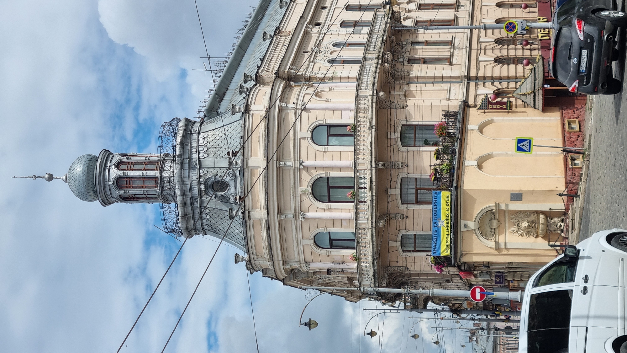 The image shows the corner of a multi-story building in Chernivtsi, Ukraine. The building has a distinctive domed tower with a spire and ornate architectural details, including columns and a decorative balcony. A banner with Ukrainian text is visible on the upper floor. In the foreground, a white van is parked on a cobblestone street. Other elements of the urban environment, such as streetlights, traffic signs, and another parked car, are also present. The sky is partly cloudy.