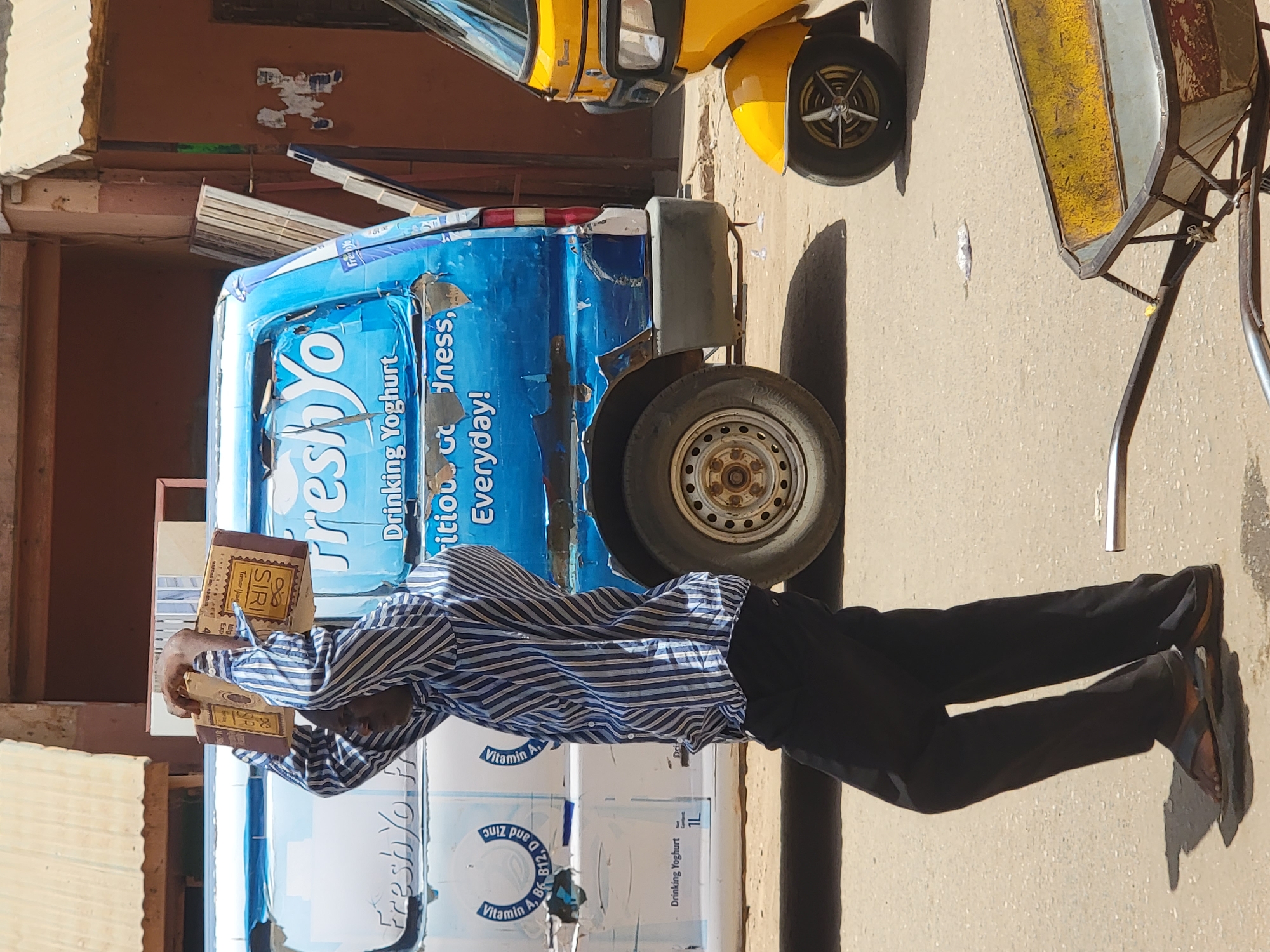 A man in a blue and white striped shirt and black pants reads two brown books in front of a Freshyo-branded truck in Maiburiji, Nigeria. The truck is parked on a paved surface and has a visible tire. A yellow vehicle, possibly a three-wheeled auto-rickshaw, is partially visible behind the truck. The environment suggests an urban street setting.