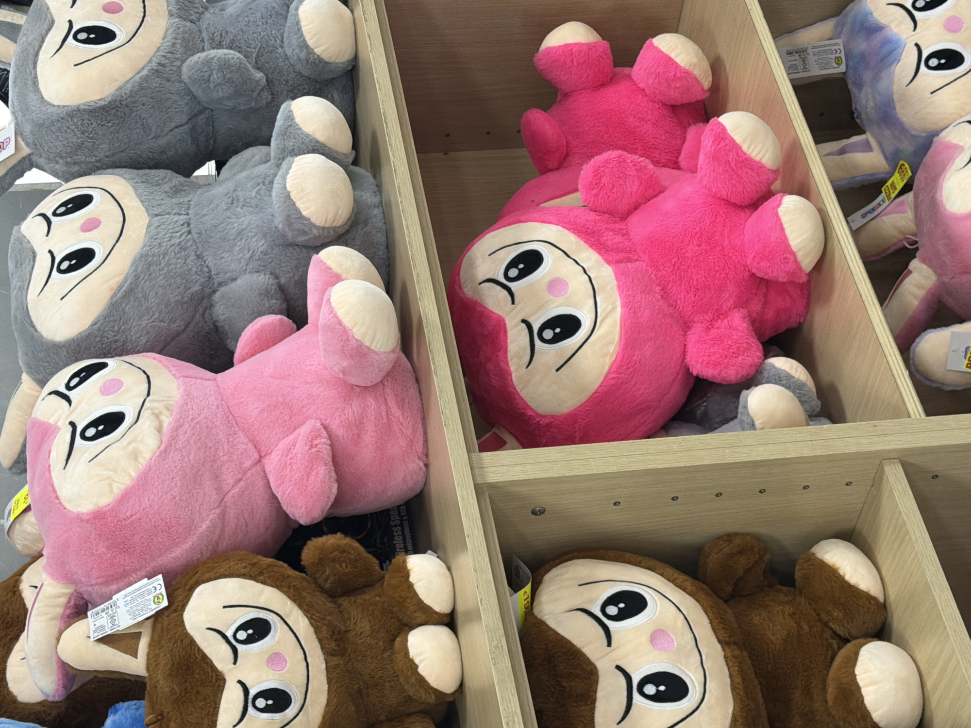 The image shows a shelf filled with stuffed animals, predominantly in shades of pink, brown, and gray. On the top shelf, from left to right, are a brown monkey-like stuffed animal, a large pink stuffed animal resembling a cartoon character with a hood, and two gray stuffed animals with similar facial features. The brown and pink stuffed animals are positioned at the front of the shelf. Below them, on the second shelf, is another large pink stuffed animal, lying down, with its face visible. To the right of this pink stuffed animal, a portion of a gray stuffed animal is visible. On the bottom shelf, partially visible, are more stuffed animals, including what appears to be a purple and white rabbit-like character. The shelves are made of light-colored wood and are part of a retail display. There are price tags and labels attached to some of the stuffed animals. The lighting suggests an indoor setting, likely a store. The image does not provide any specific information or visual cues to determine its location in Due Carrare, Italy.