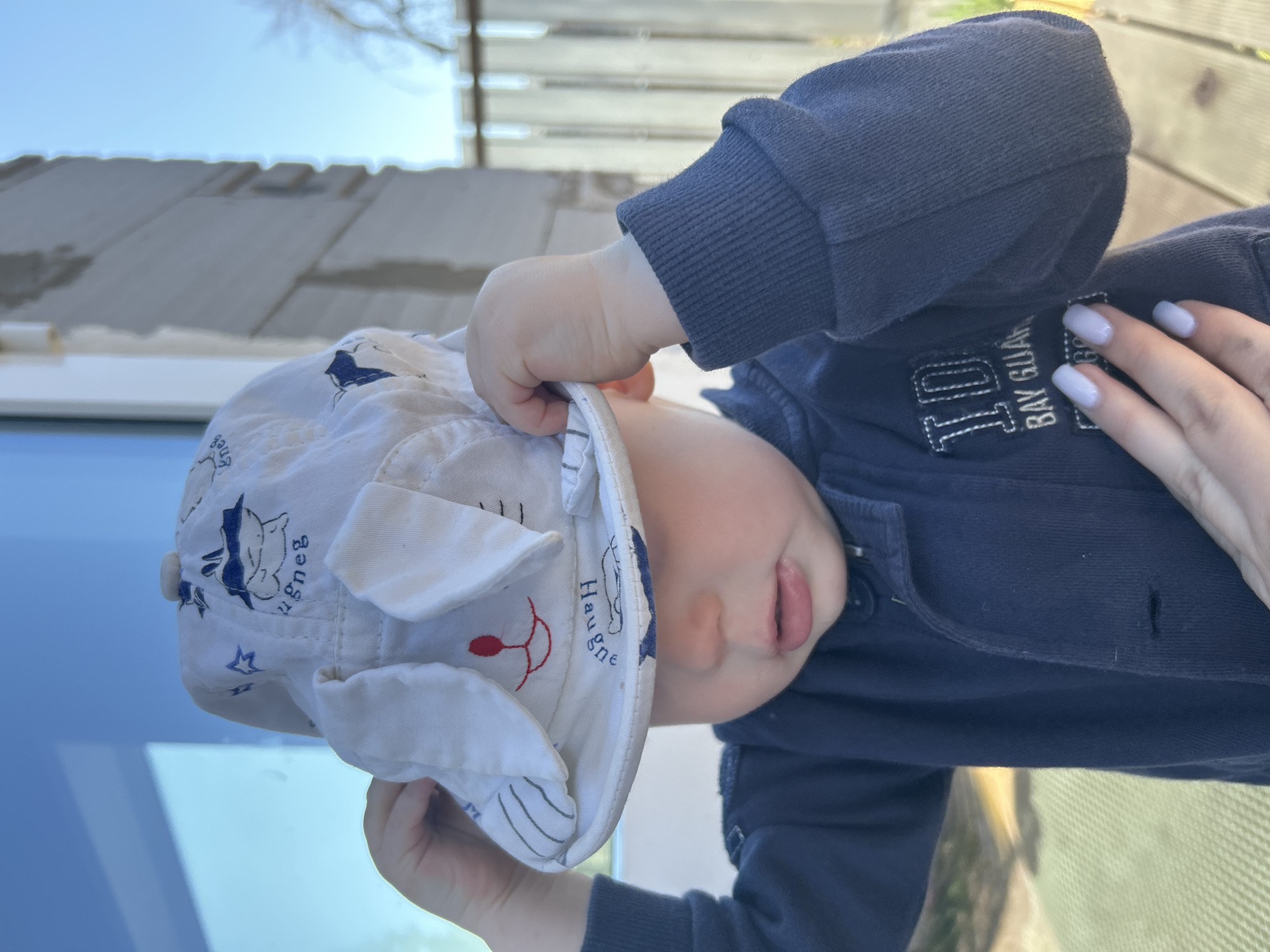 A young child wearing a blue jacket and a white hat with bunny ears is outdoors. The child is holding the hat with one hand, while the other hand is visible with light blue painted nails. The background includes a partially constructed building and foliage. The location mentioned is Chaikovsky, Russia.