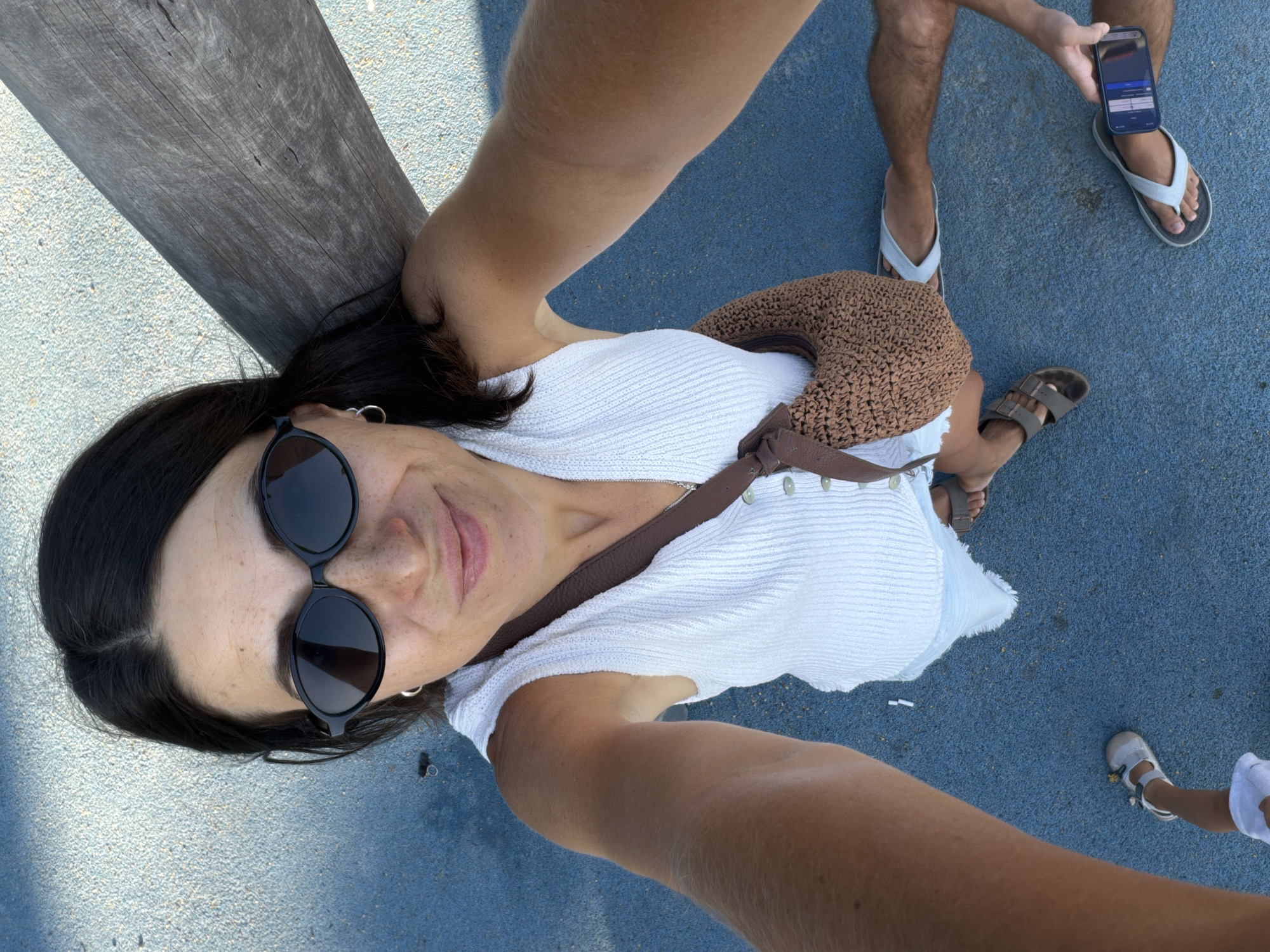 A woman with dark hair and sunglasses is taking a selfie outdoors, leaning against a wooden beam. She is wearing a white sleeveless top and a brown woven shoulder bag. She is on a blue surface, possibly a playground or a public area. In the lower part of the image, a pair of feet wearing sandals are visible, and another person is holding a smartphone. The overall impression is a casual, sunny day. The location is the city of Budva, Montenegro.