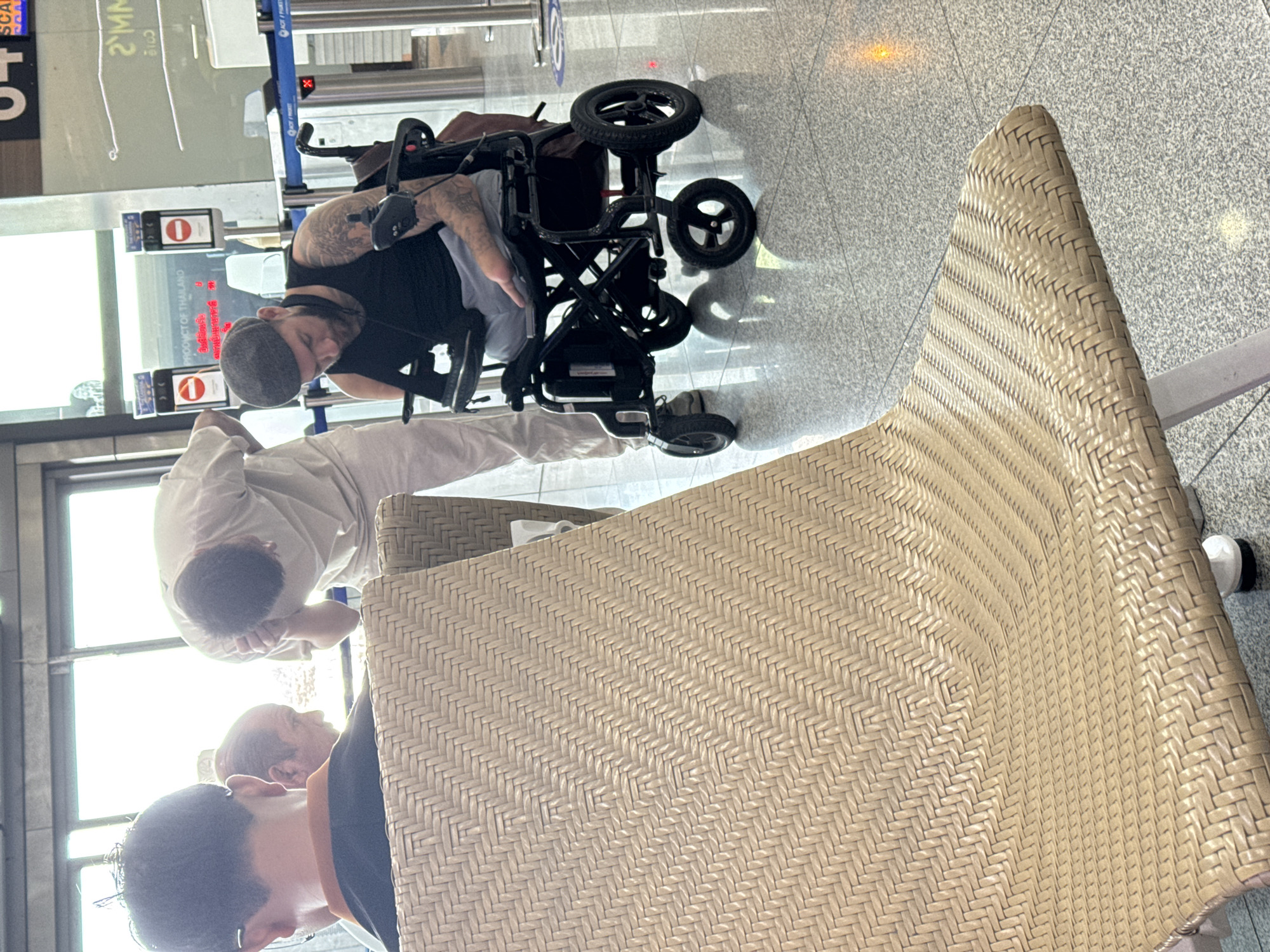 In Mai Khao, Thailand, an individual with tattoos on their arms sits in a black electric wheelchair, wearing a black tank top and a grey flat cap, and appears to be looking down at a device in their lap. To the left, another person in a black shirt with an orange collar is seated, with the back of their head visible, facing away from the camera. Behind them, a third individual in a light-colored uniform is bent over, with their head in their hands, possibly attending to something on the floor. The scene takes place in what appears to be an airport terminal, indicated by the presence of a gate or checkpoint with red "stop" signs and stanchions. A woven chair occupies the foreground, partially obscuring the view of the people and the environment. The floor is a polished, reflective tile.
