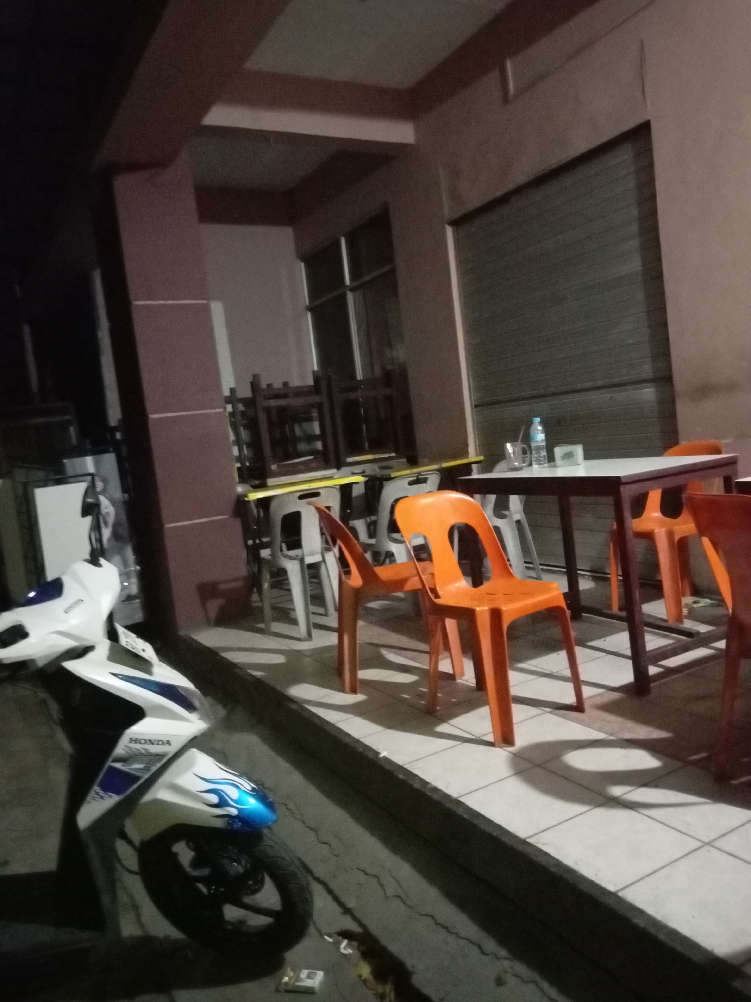 A horizontal, dimly lit, eye-level outdoor shot shows an arrangement of tables and chairs outside a commercial establishment. The scene is likely a food stall or small restaurant in Batam, Indonesia. In the foreground, a white and blue Honda scooter is parked on a sidewalk. Beyond the scooter, several tables with white tops and dark frames are set up, surrounded by various chairs. Some chairs are stacked, while others are scattered around the tables. Among the chairs are bright orange plastic ones and white plastic ones. On one of the tables, a bottle of water, a glass, and a small white container are visible. In the background, a gray rolling shutter door is closed, indicating the establishment is not open for business. The area appears to be a public space, possibly a sidewalk or an open-air seating area.