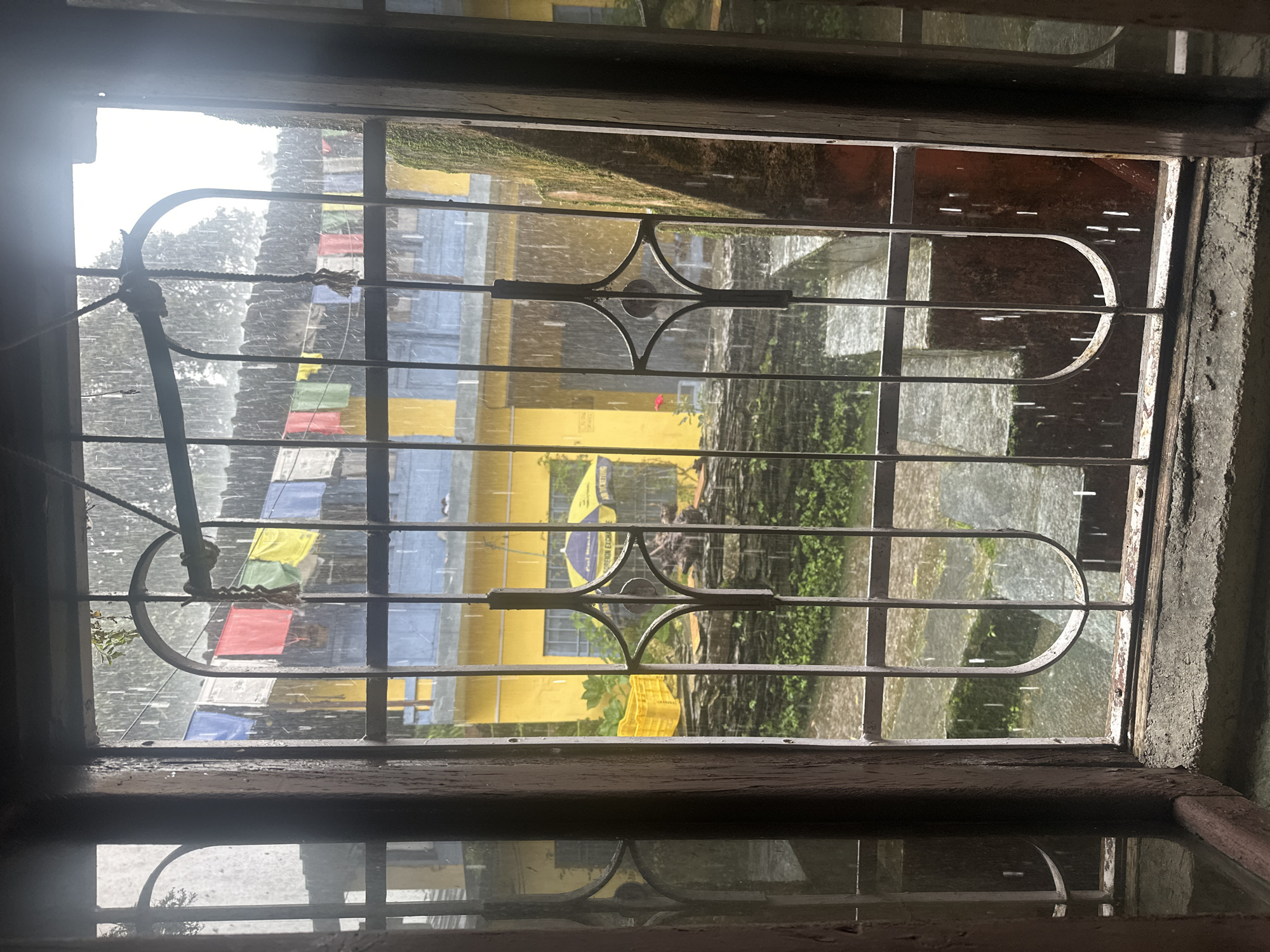 The image is an interior view through a barred window looking out at a rainy day in Haini, India. Prayer flags in various colors (blue, red, yellow, green, white) are strung across the scene above a yellow building with blue trim. A yellow umbrella is visible in front of the building. There are also several yellow crates and some greenery outside. The rain is clearly visible, streaking down the windowpane and the exterior. The window frame is made of dark wood and appears old and weathered. The bars are made of metal with decorative scrollwork. The ground outside appears wet and covered in mossy stones.