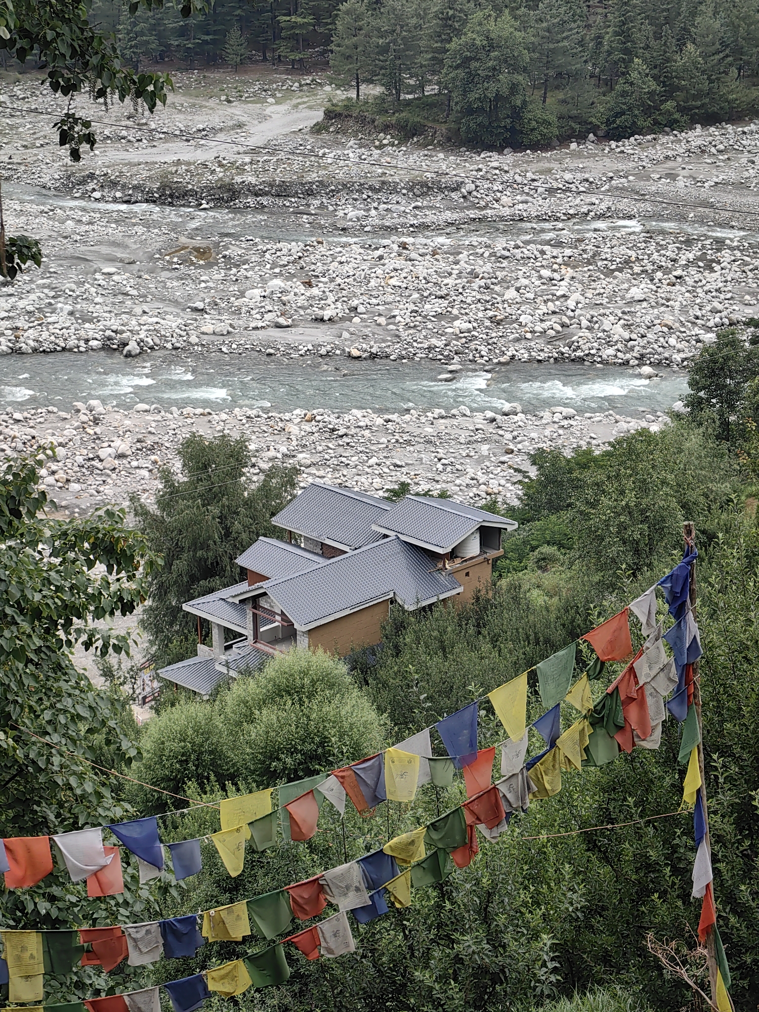 The image depicts a mountainous landscape in Vashisht, India, characterized by a wide, stony riverbed with a flowing river. The river is shallow with visible currents and is bordered by numerous grey and white stones. In the mid-ground, a multi-level building with dark grey tiled roofs is nestled amidst dense green foliage and trees. Prayer flags in various colors, including blue, white, red, green, and yellow, are strung across the foreground, adding a vibrant touch to the scene. The flags are suspended on strings, creating a decorative element that partially obscures the view of the building and the river. The background is dominated by a dense forest of pine trees, suggesting a remote or natural setting. The overall impression is one of a serene, natural environment with a touch of cultural influence from the prayer flags.