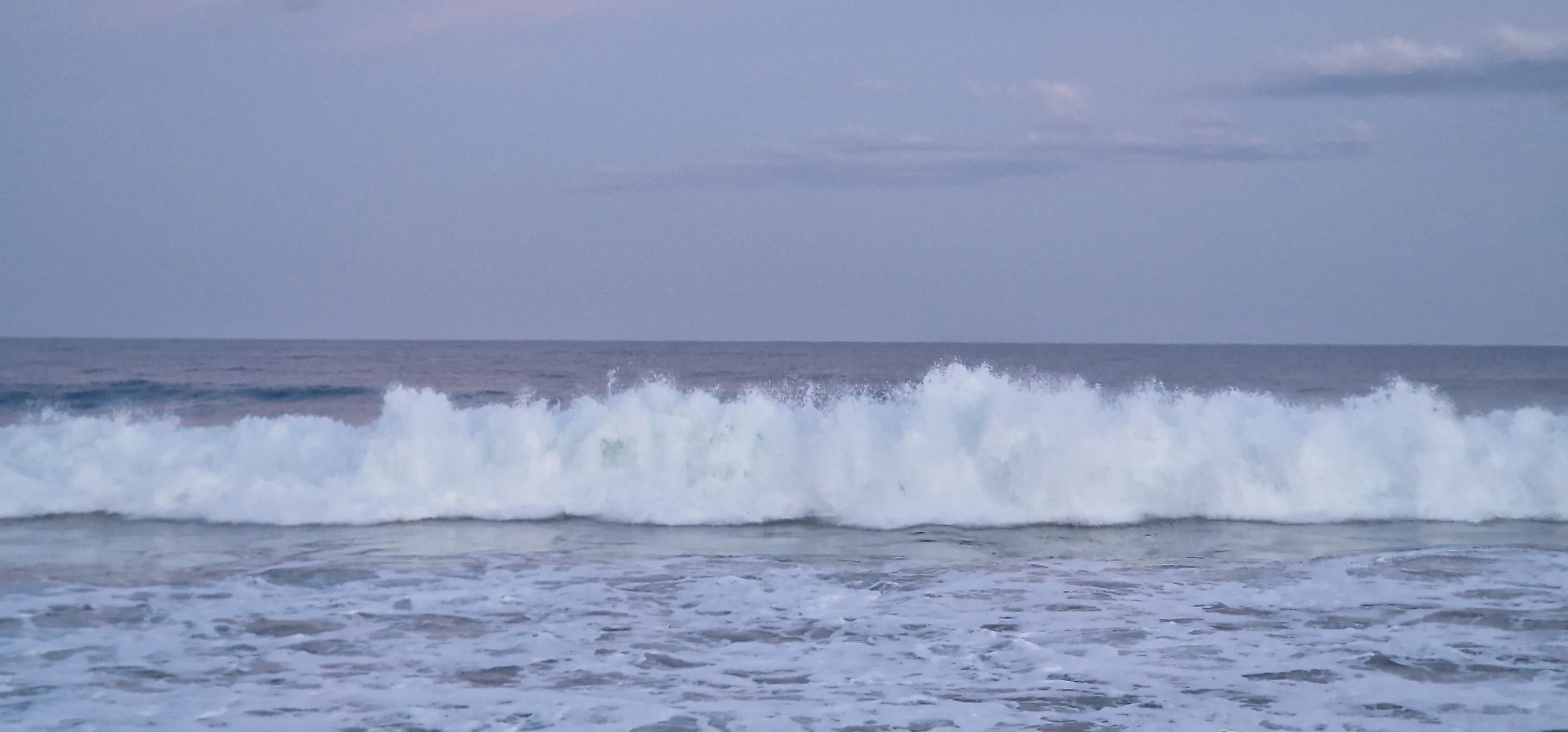 The image displays a serene yet powerful ocean scene, centered on a large wave breaking. The main subject is a substantial white wave, horizontally bisecting the frame in the mid-ground, showing turbulent foam and spray as it crashes. In the foreground, the water is a mix of white froth and darker blue-gray, indicating receding or previously broken waves. Beyond the breaking wave, the calm, deep blue-gray ocean stretches to the horizon.
The sky occupies the upper half of the image and is a soft, muted lavender-blue-gray, suggesting either dawn or dusk due to the diffused light and lack of strong shadows. A few faint, wispy clouds are discernible in the upper sky. The color palette is dominated by various shades of blue, gray, and white, creating a tranquil and natural atmosphere. No people, objects, or text are visible, emphasizing the raw, untamed beauty of the sea. The setting is a coastline, characteristic of a beach in Pottuvil, Sri Lanka.