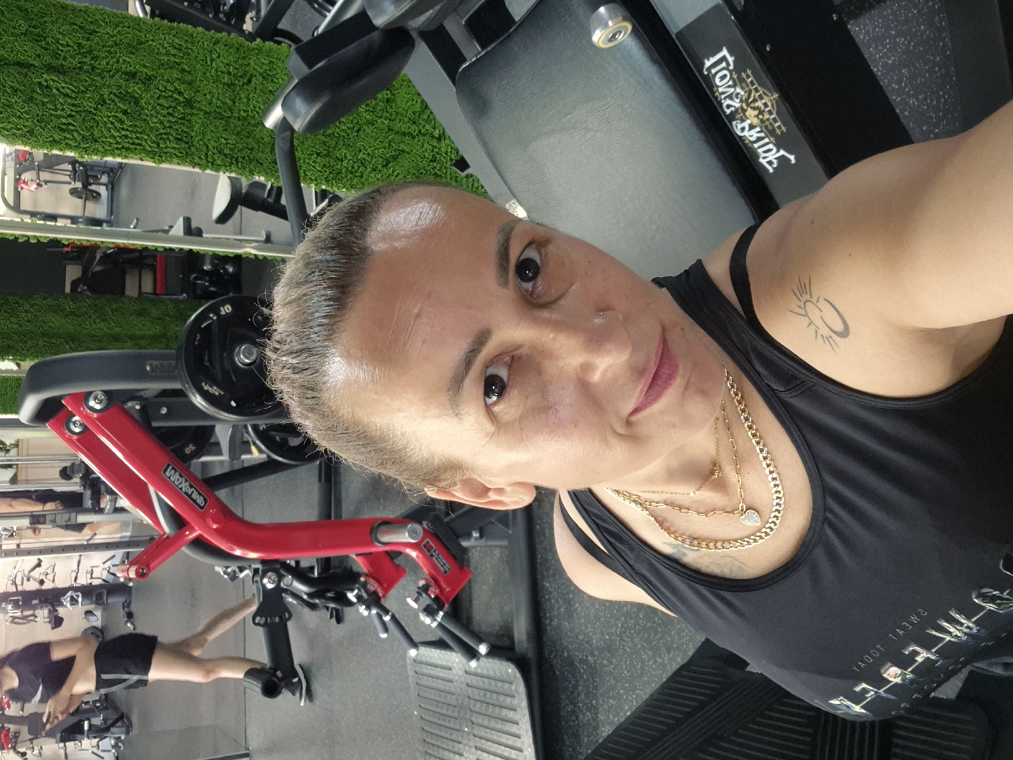 A woman takes a selfie in a gym, her head and upper body filling the foreground. She wears a black tank top with gold layered necklaces and a small tattoo of a crescent moon with a sun on her left shoulder. In the background, gym equipment is visible, including a red leg press machine and other weight machines. A section of artificial green turf is on the right. The lighting suggests an indoor environment.