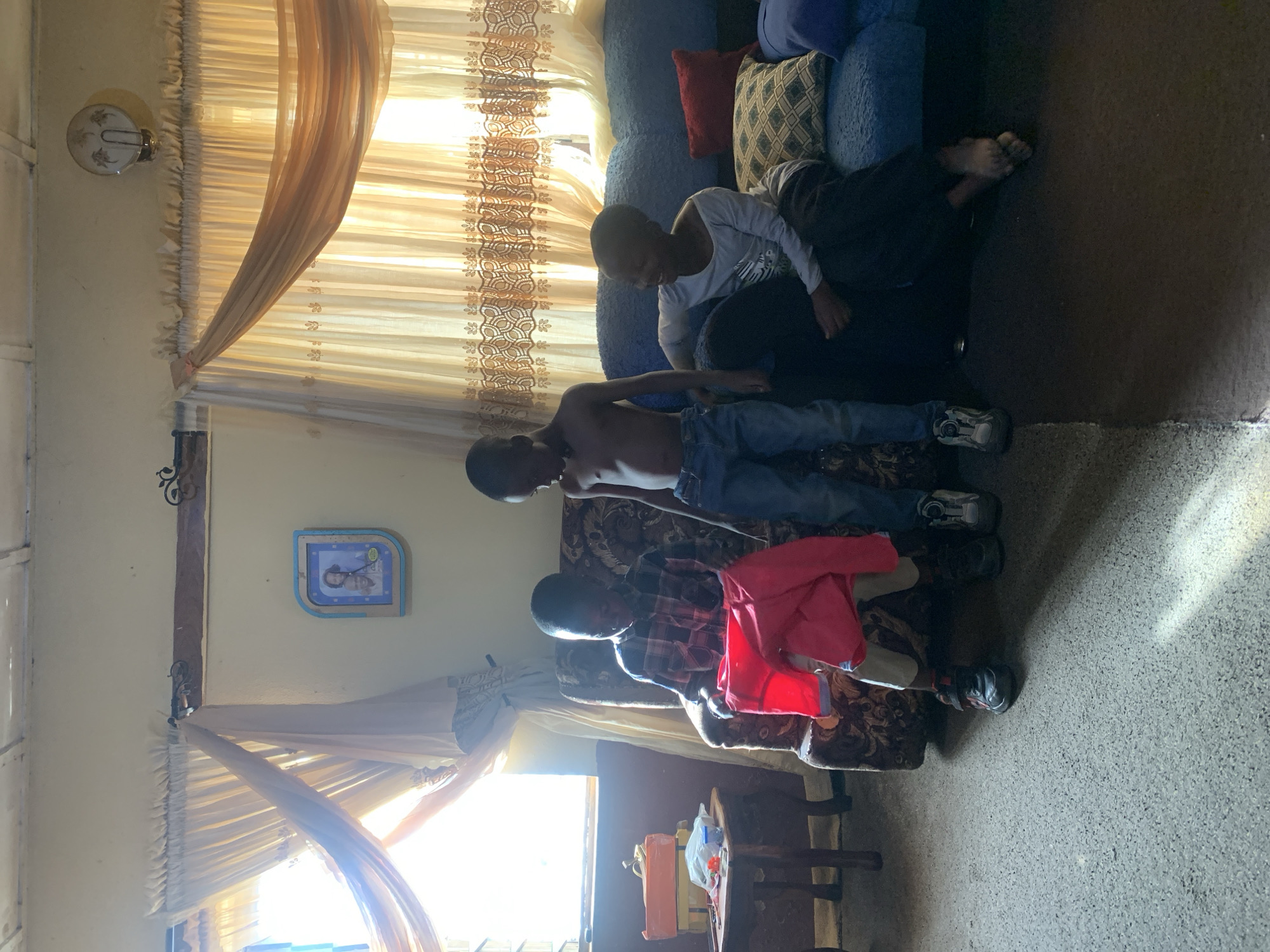 In Rafin Bauna, Nigeria, several individuals are gathered indoors. Two boys are visible on a floral patterned couch with a red jacket resting nearby. A third boy sits on a blue, pillowy couch. The setting features patterned curtains and walls decorated with what appears to be a clock. A window is visible in the background, adding to the indoor environment. The floor appears to be bare.