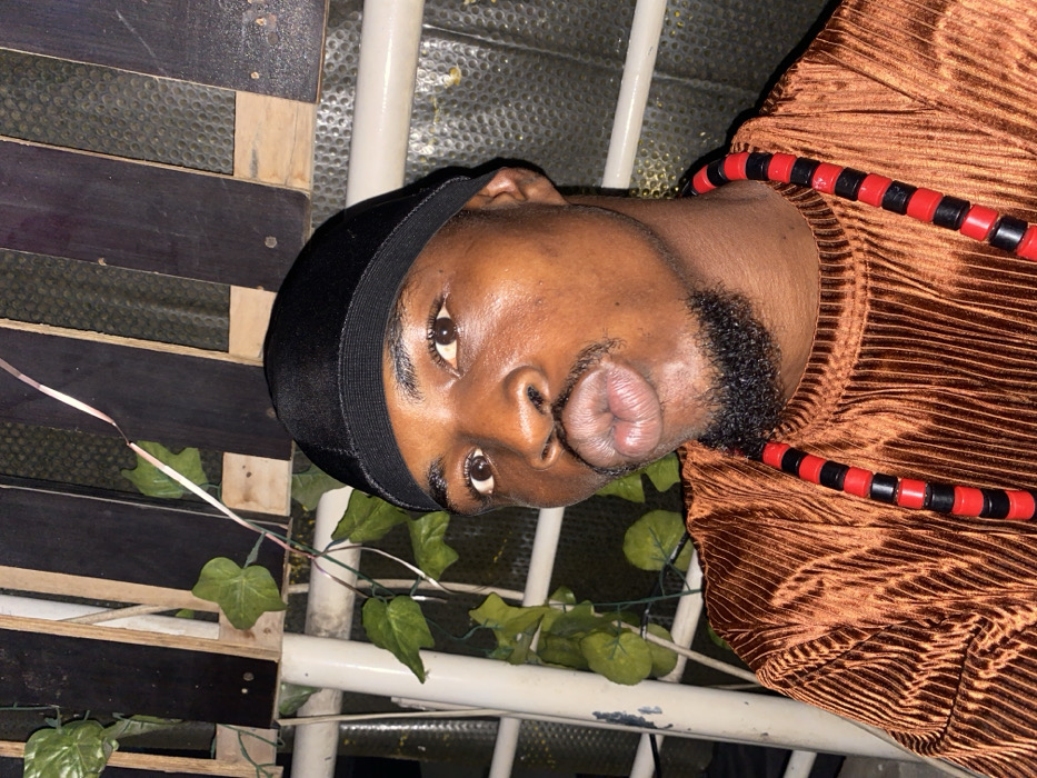 A close-up shot shows a person wearing a black durag, a brown, textured, long-sleeved top, and a red and black beaded necklace. The person has dark skin, a beard, and is puckering their lips. Behind the person are plants with green leaves and a wooden pallet. White metal bars are also visible. The image is shot from a low angle and is slightly rotated to the side.