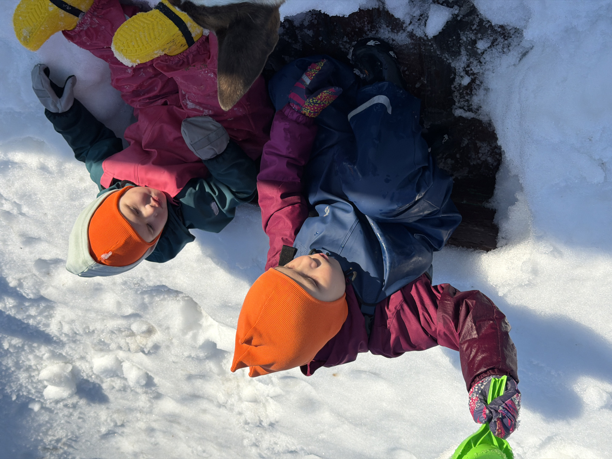 Two children are lying on their backs in the snow in an outdoor environment. The child on the left is wearing a pink snowsuit, yellow boots, grey gloves, and an orange and light green hat. The child on the right is wearing a maroon and blue snowsuit, black boots, multi-colored gloves, and an orange hat. The child on the right is holding a green plastic toy. The ground is covered in snow, with a dark, possibly muddy or wooden, area behind the children. The location is presumed to be the city of Chaikovsky, Russia.