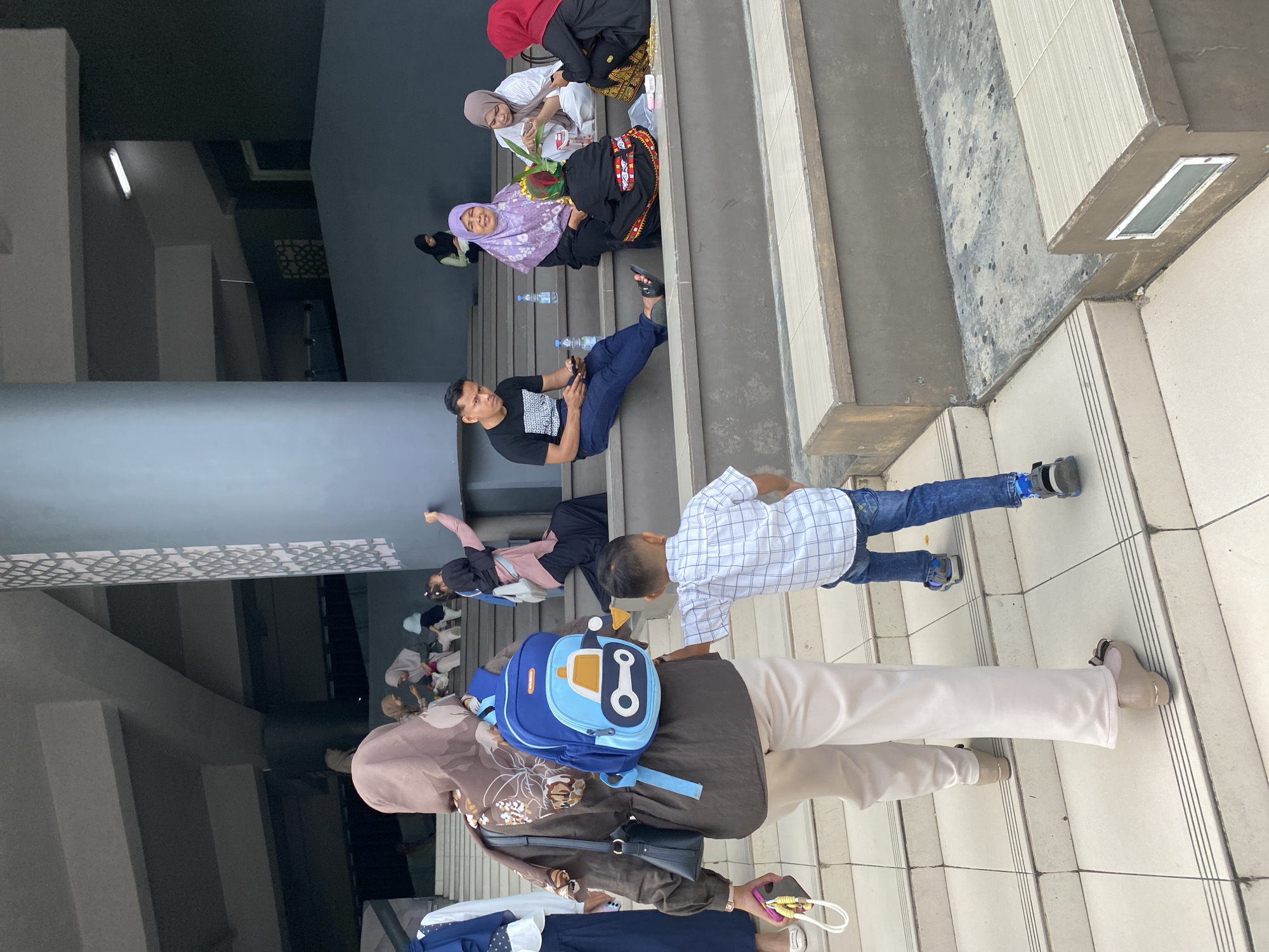 In Banda Aceh, Indonesia, a group of people are gathered on a wide, tiered outdoor staircase, likely part of a public building or complex. The architecture features a modern, minimalist design with grey concrete and tiled steps.
In the foreground, a woman wearing a brown hijab and a long-sleeved brown shirt is walking up the stairs, carrying a black handbag and a small blue backpack with a cartoon car design. A young boy in a white checkered shirt and blue jeans walks slightly ahead of her, also ascending the stairs.
Further up the staircase, several individuals are seated. On the left, a man in a black t-shirt and blue jeans is looking at his phone. To his right, a woman in a purple hijab is smiling, and next to her, another woman in a light-colored hijab is holding something in her hands. A person in a black outfit with colorful accents is seated in front of them. On the far right, a woman with a red hijab is also seated.
Other individuals can be seen further up and in the background, suggesting a gathering or a public event. The overall scene depicts a casual and communal atmosphere on a bright day.