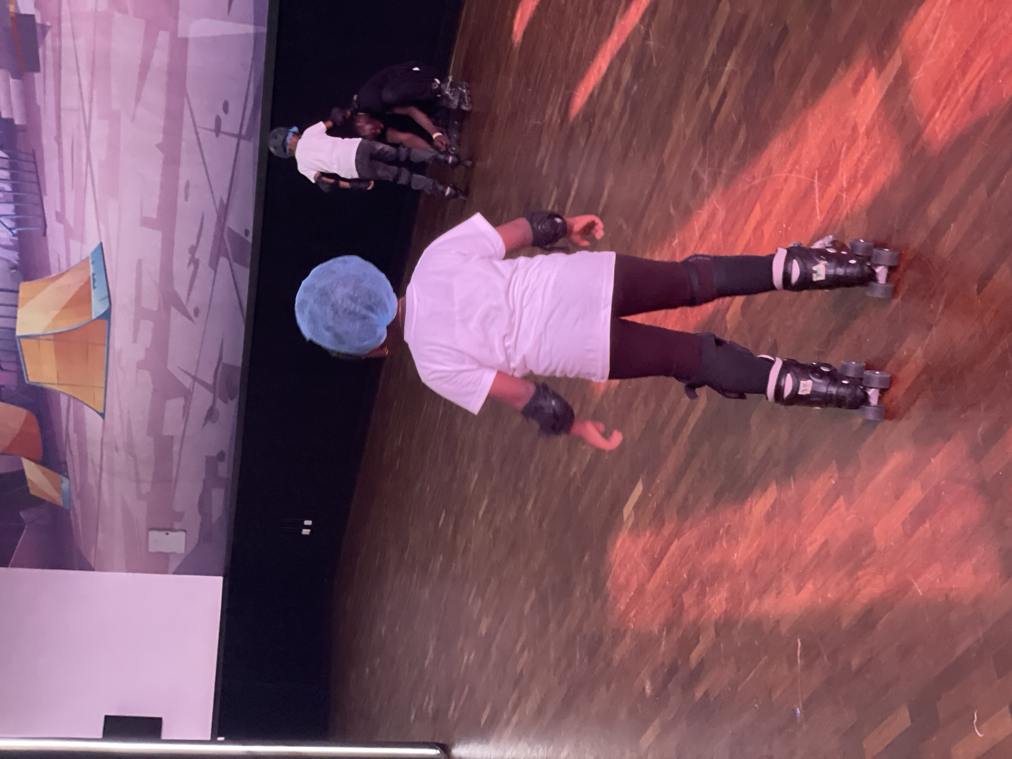 A person with a blue hair covering is wearing roller skates, knee pads, and wrist guards on a hardwood floor. Three people in similar safety gear are in the background. A painted wall is on the left. The location is not discernible as Moba, Nigeria.