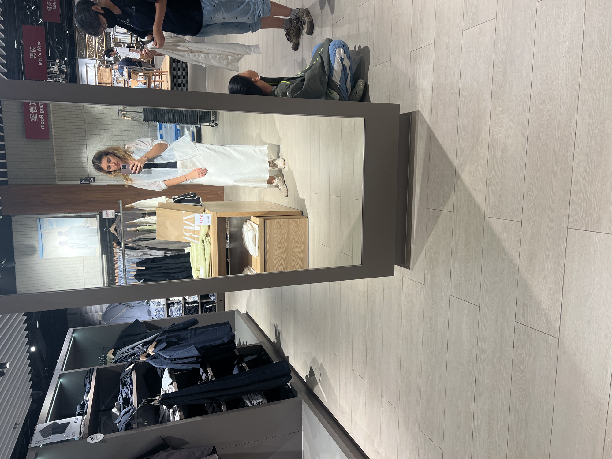 A wide-angle, eye-level shot captures a brightly lit retail store, likely a clothing boutique, with light wood-look flooring. In the foreground, a large floor-to-ceiling mirror reflects the scene. A woman with blonde hair, wearing a white button-down shirt over a black top and a long white skirt, stands in front of the mirror, holding her phone to take a selfie. Her light brown shoes have a casual lace-up style. To her right, a woman with dark hair and a black mask covering her face is crouched down and looking up towards the mirror, holding a white garment. Her attire includes a grey hoodie with blue and white accents and blue sweatpants with white stripes. In the background, male shoppers are visible, one wearing a black t-shirt and shorts and another in a white t-shirt and dark pants. Clothing racks with various garments, predominantly in dark colors like navy and black, are organized on shelving units to the left and in the background. A wooden display table with folded clothes and a paper bag is positioned to the right of the mirror. Overhead lights illuminate the store. The signage above the mirror is in Chinese characters and English, with "Men's Wear" visible. The overall impression is that of a contemporary, well-organized retail space. The presence of Chinese characters on the signage suggests the location is in Hong Kong, China.