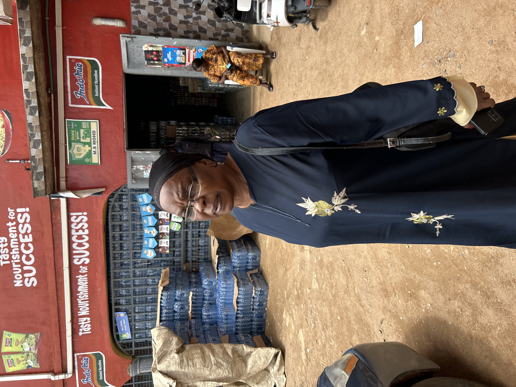 In the city of Jos, Nigeria, a woman wearing a dark blue abaya with embroidered gold flowers stands in the foreground, looking to her right. She has glasses and a black headscarf. Behind her, a store is visible with shelves stocked with bottled water and other products. A sign above the store entrance reads "Tasty Nourishment for SUCCESS!" with the Indomie logo. Further inside the store, another woman in an orange patterned outfit is seated. A motorcycle is parked to the right of the store. The ground is dusty, and a dark-colored car is partially visible on the left side of the image.