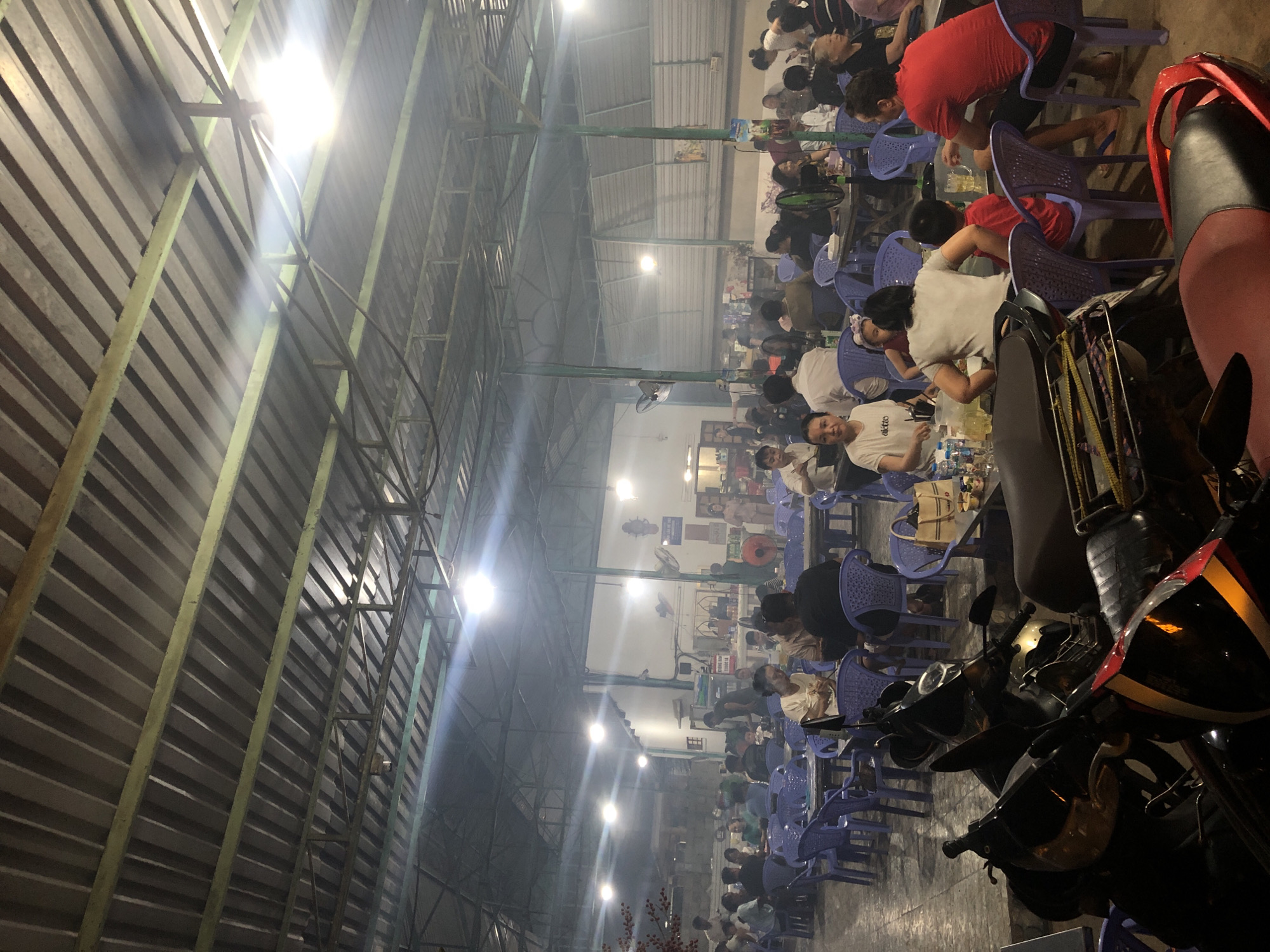 The image depicts a busy outdoor dining area, likely a restaurant or street food stall, in Phu Thuy Ward, Vietnam. The setting is covered by a corrugated metal roof supported by metal beams, indicating an open-air structure. Numerous people are seated at tables, engaged in dining and socializing. The atmosphere appears lively, with a moderate amount of smoke or haze present, possibly from cooking or ambient conditions.
The subjects are primarily Vietnamese people of various ages, dressed casually. They are seated on blue plastic chairs around tables, many of which are laden with food and drinks. The activity is clearly a communal meal or gathering. In the foreground, several motorcycles are parked, suggesting this is a common mode of transport for patrons.
Notable details include the time of day, which appears to be evening or night due to the artificial lighting, with bright overhead lights illuminating the scene. The weather is not explicitly discernible, but the covered setting suggests it might be for protection from sun or rain. The presence of multiple fans indicates a potentially warm climate. The overall mood seems to be relaxed and convivial. No discernible text is visible in the image.