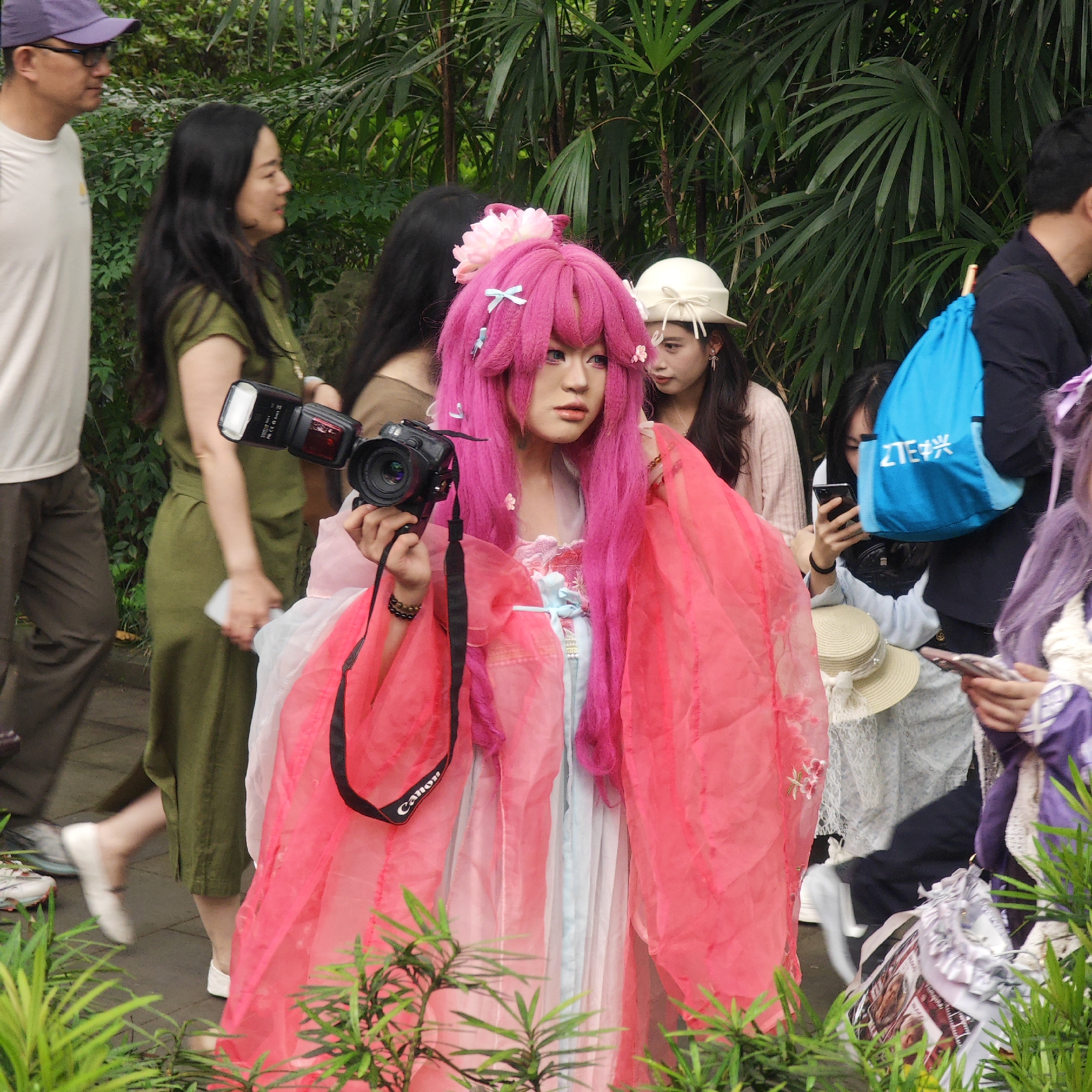 The image, taken in Yuzhong District, China, features an individual dressed in a pink cosplay outfit, complete with a pink wig adorned with flowers and ribbons. The person is holding a Canon camera with a flash attachment. In the background, several other people are visible. One woman in a green jumpsuit walks past, while others are standing or sitting nearby, some holding phones and a blue backpack. The setting appears to be an outdoor park or garden area with lush greenery in the background and foreground. The image captures a gathering of individuals, possibly for a cosplay event or photoshoot, in a public park setting.