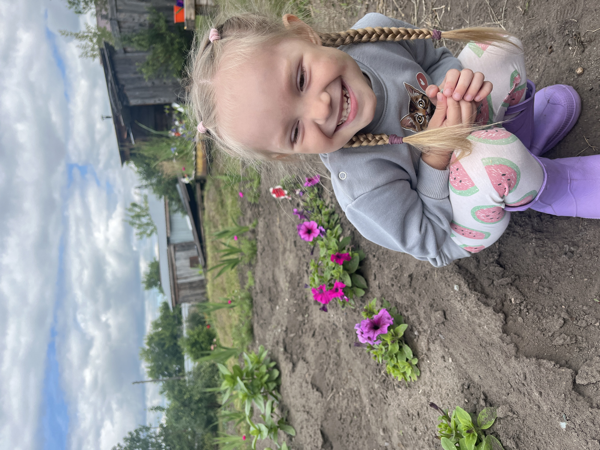 A young girl with blonde hair in two braids, wearing a grey sweatshirt with a cat graphic and white pants with a watermelon print, crouches in a garden. She is smiling broadly, with her eyes closed. In the background, there are several small buildings, trees, and a cloudy sky. The garden appears to be in a rural setting, likely in or near the village of Сухринское, Russia. The ground is dark soil, with some green plants and pink and purple flowers visible. The girl is wearing purple boots.