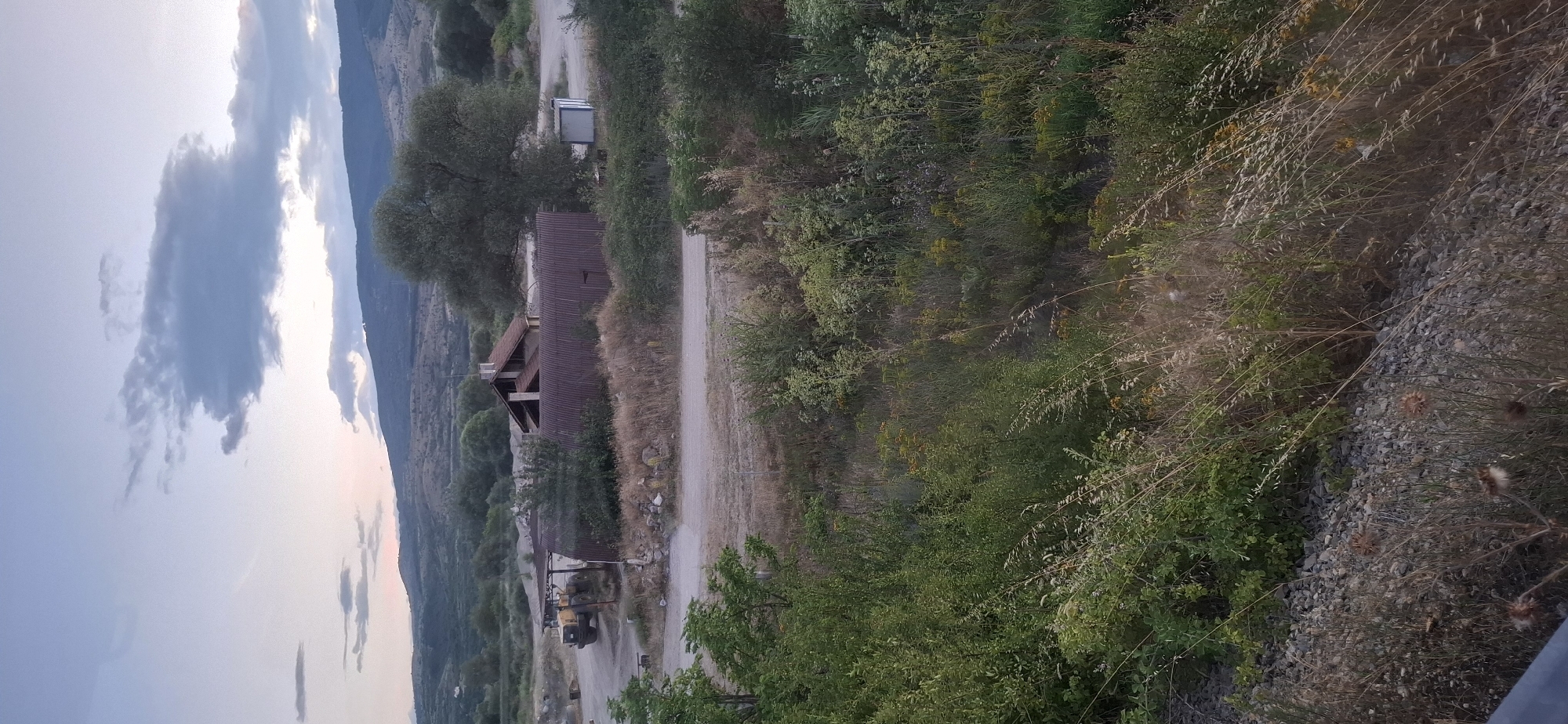 In Kocherinovo, Bulgaria, the image captures a rural landscape at dusk, featuring a cluster of buildings and natural elements. A yellow loader, a heavy construction vehicle, is parked near a covered structure, suggesting industrial or agricultural activity. Adjacent to it stands a building with a red, corrugated metal roof, appearing weathered and aged. Further to the right, a smaller, lighter-colored structure with a blue-tinted window and white trim is visible. The area is surrounded by abundant vegetation, including dense bushes, tall grasses, and scattered trees, with a large, mature tree dominating the mid-ground. In the background, rolling hills are silhouetted against a sky filled with soft, scattered clouds, illuminated by the fading light of sunset. The terrain appears uneven, with a gravelly ground surface around the structures.
