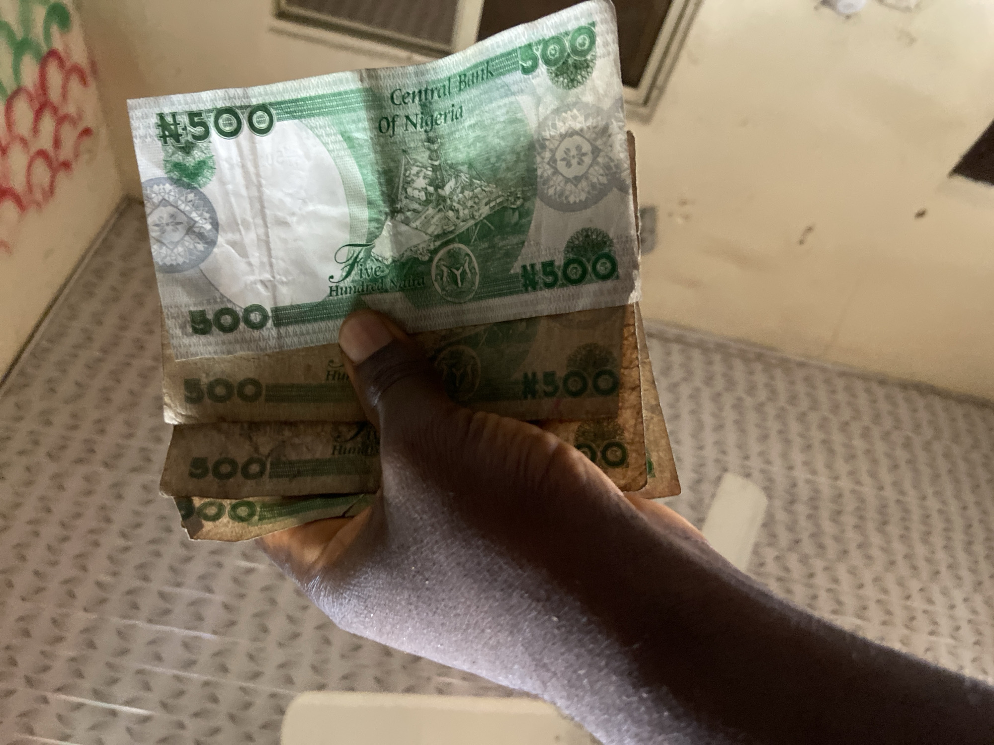 A dark-skinned hand is prominently displayed, holding a stack of Nigerian 500 Naira banknotes. The top banknote is clearly visible, featuring the denomination "#500" and "500" in various locations. The text "Central Bank Of Nigeria" and "Five Hundred Naira" are distinctly legible on the note. Its design includes an illustration that appears to be a ship or oil rig, along with complex patterns and a large blank watermark area. Several other 500 Naira notes are partially visible beneath the top one.
The scene is set indoors, with a light-colored, possibly tiled or patterned floor visible below and to the left of the hand. A wall is also present in the background, adorned with some red and green circular markings or doodles. The lighting is diffused, suggesting an interior space without direct natural light indicating a specific time of day. The focus is tightly on the hand and the currency it holds, providing a clear view of the Nigerian banknotes. The location is Mararaba, Nigeria.