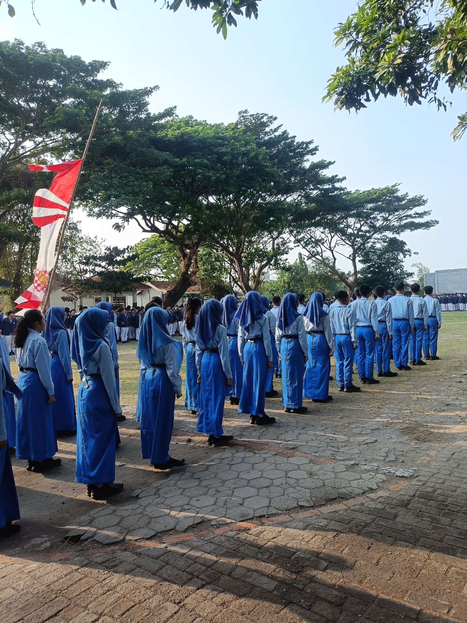 The image depicts a group of students in uniform standing in formation outdoors, likely in a schoolyard or parade ground, in Kediri City, Indonesia. The students, predominantly female, are wearing light blue shirts, blue skirts or pants, and headscarves, suggesting a school uniform. Some students are wearing white shirts with blue pants and headscarves. A red and white flag, possibly the Indonesian flag, is visible on a bamboo pole in the background, partially obscured by trees. The ground is paved with interlocking bricks. The students appear to be at attention or awaiting instructions. The environment suggests a daytime scene with moderate sunlight and shadows. In the distance, a building with a tiled roof is visible, along with lush green trees. The overall impression is of an organized gathering, possibly a flag-raising ceremony or a school assembly.